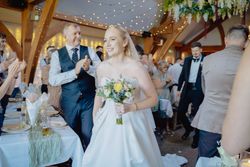 Smiling bride in a white wedding dress holding a bouquet, walking into a wedding reception with guests applauding.