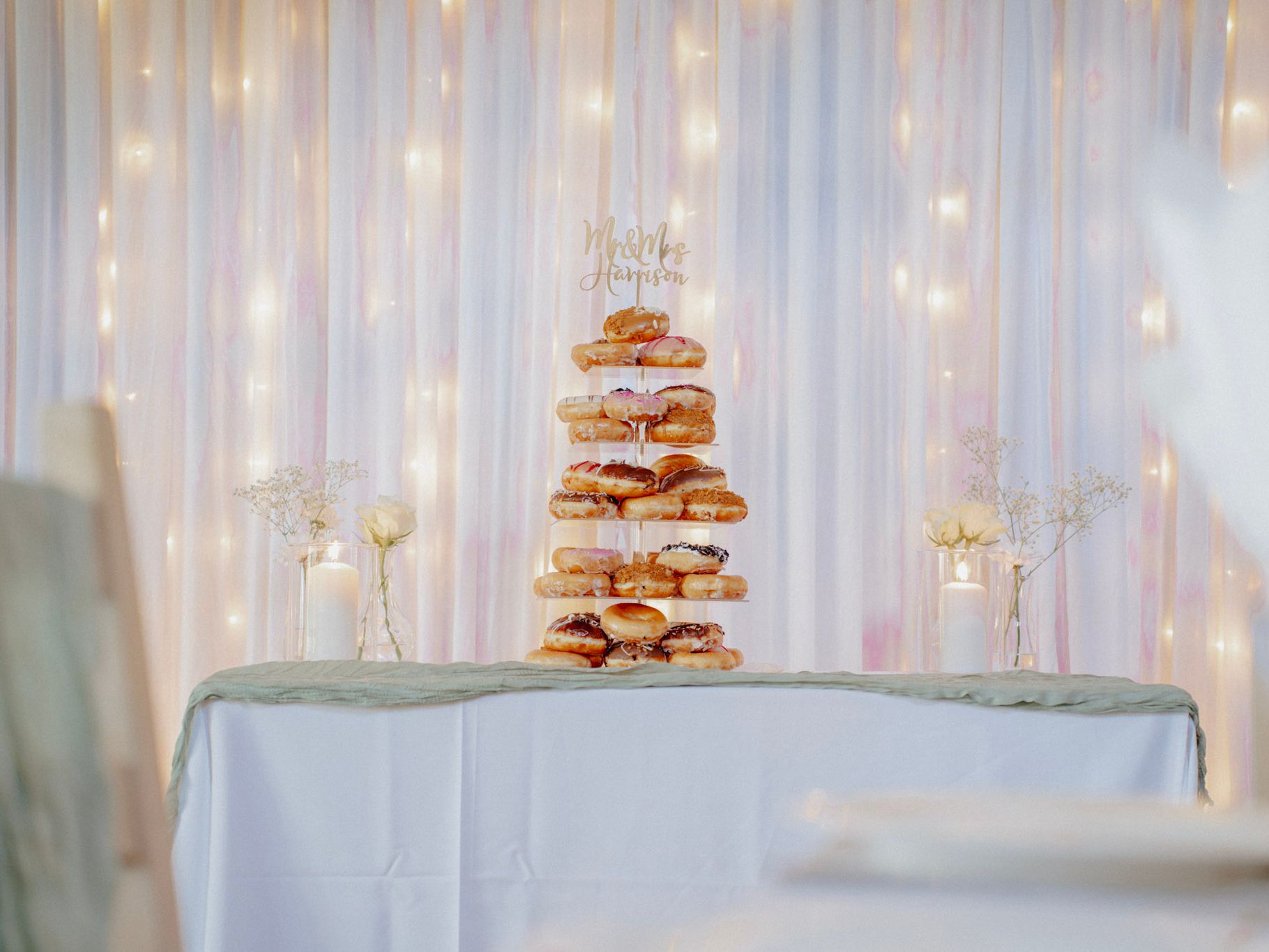 A tiered donut tower with a 'Mr & Mrs Harrison' topper on a decorated table with white flowers and candles in front of a backdrop of fairy lights and white drapery.