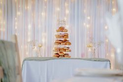A tiered donut tower with a 'Mr & Mrs Harrison' topper on a decorated table with white flowers and candles in front of a backdrop of fairy lights and white drapery.