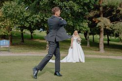 A groom in a suit swings a golf club while a bride in a white dress stands smiling behind him on a grassy area with trees.