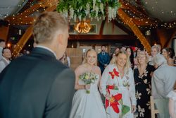 Bride walking down the aisle accompanied by a smiling woman, surrounded by guests in a decorated wedding venue.