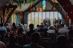 Wedding ceremony indoors with guests seated and groomsmen standing at the front, large windows and wooden beams in the background.