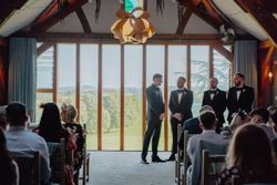 A group of groomsmen in tuxedos standing at the front of a ceremony hall with guests seated, backed by large windows with a scenic view.
