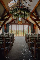 Indoor wedding ceremony setup with wooden chairs, green drapery, flower petals on the aisle, and large windows overlooking a scenic landscape.