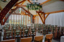 Rustic indoor wedding ceremony setup with wooden chairs, green sashes, fairy lights, and a floral chandelier.