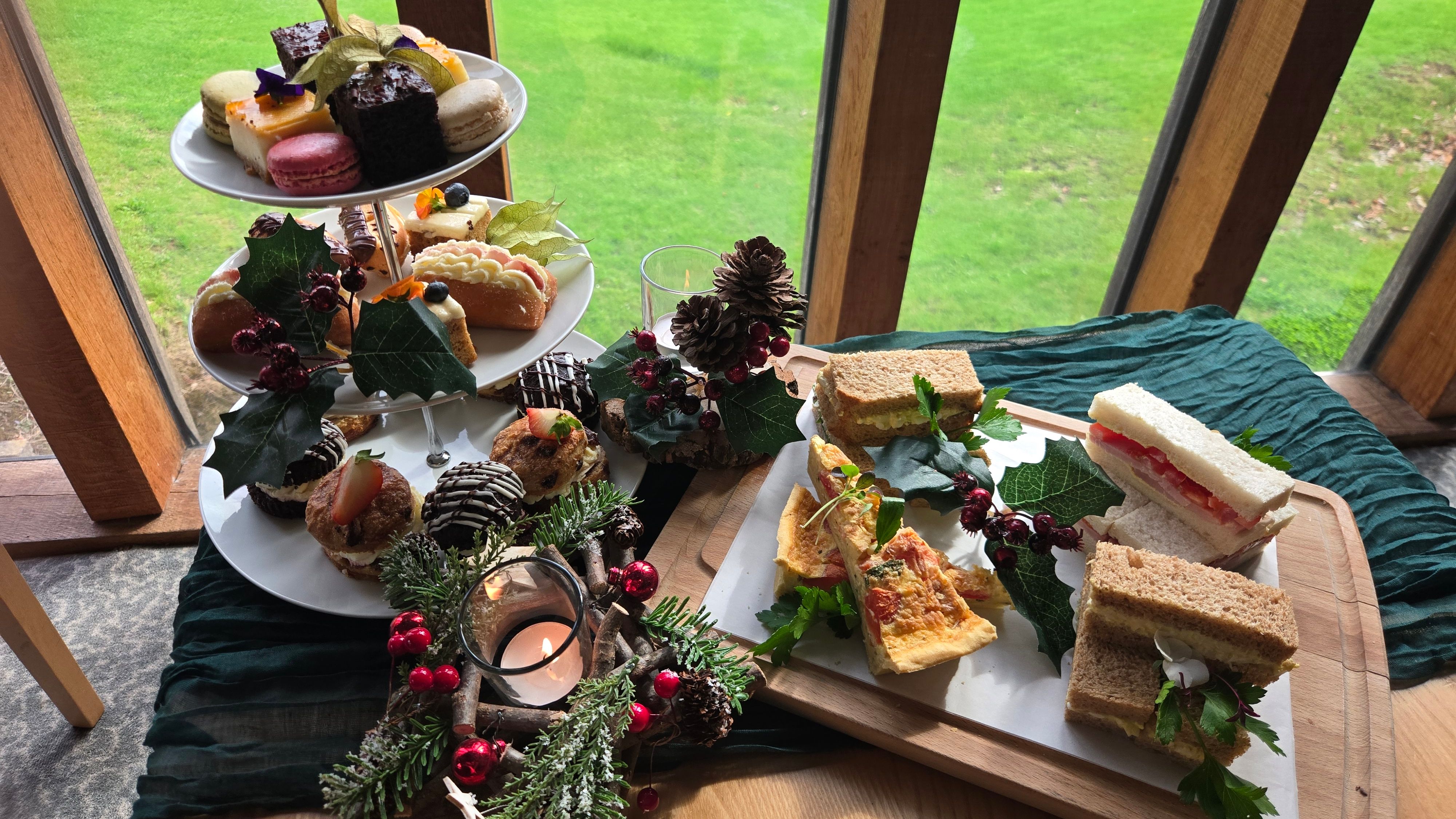 Traditional afternoon tea set with assorted finger sandwiches and desserts by a window overlooking a green lawn.