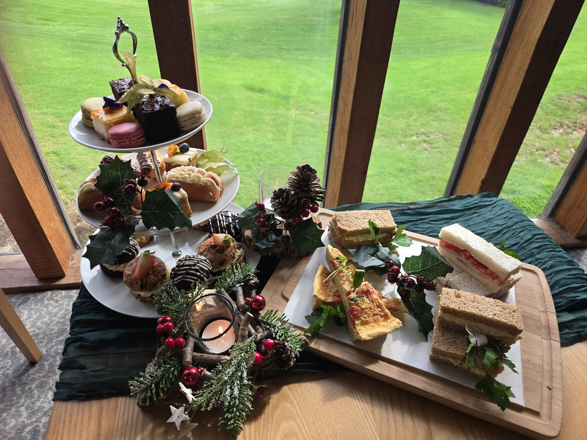 Afternoon tea set with assorted pastries, sandwiches, and festive decorations on a wooden table by a window overlooking green grass.