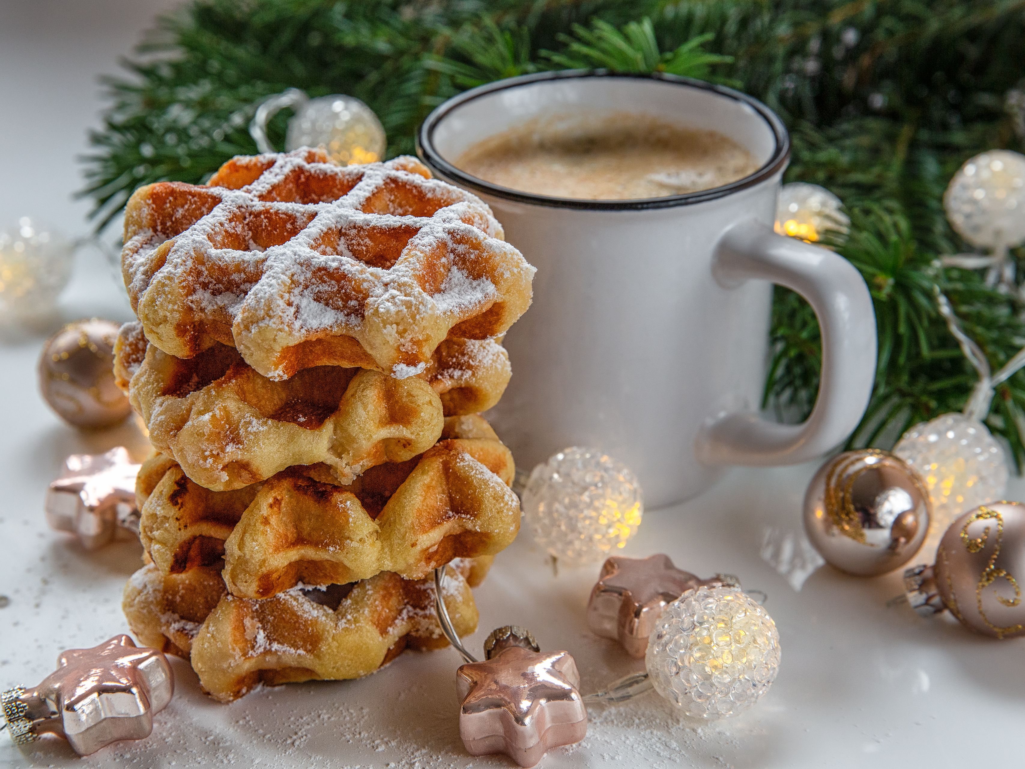 Stack of waffles topped with powdered sugar next to a mug of coffee, surrounded by Christmas decorations and greenery.