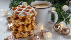 Stack of waffles topped with powdered sugar next to a mug of coffee, surrounded by Christmas decorations and greenery.