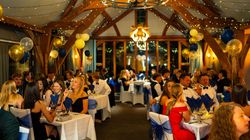 Elegant formal dinner event with people dressed up, sitting at decorated tables with blue and gold balloons and fairy lights.