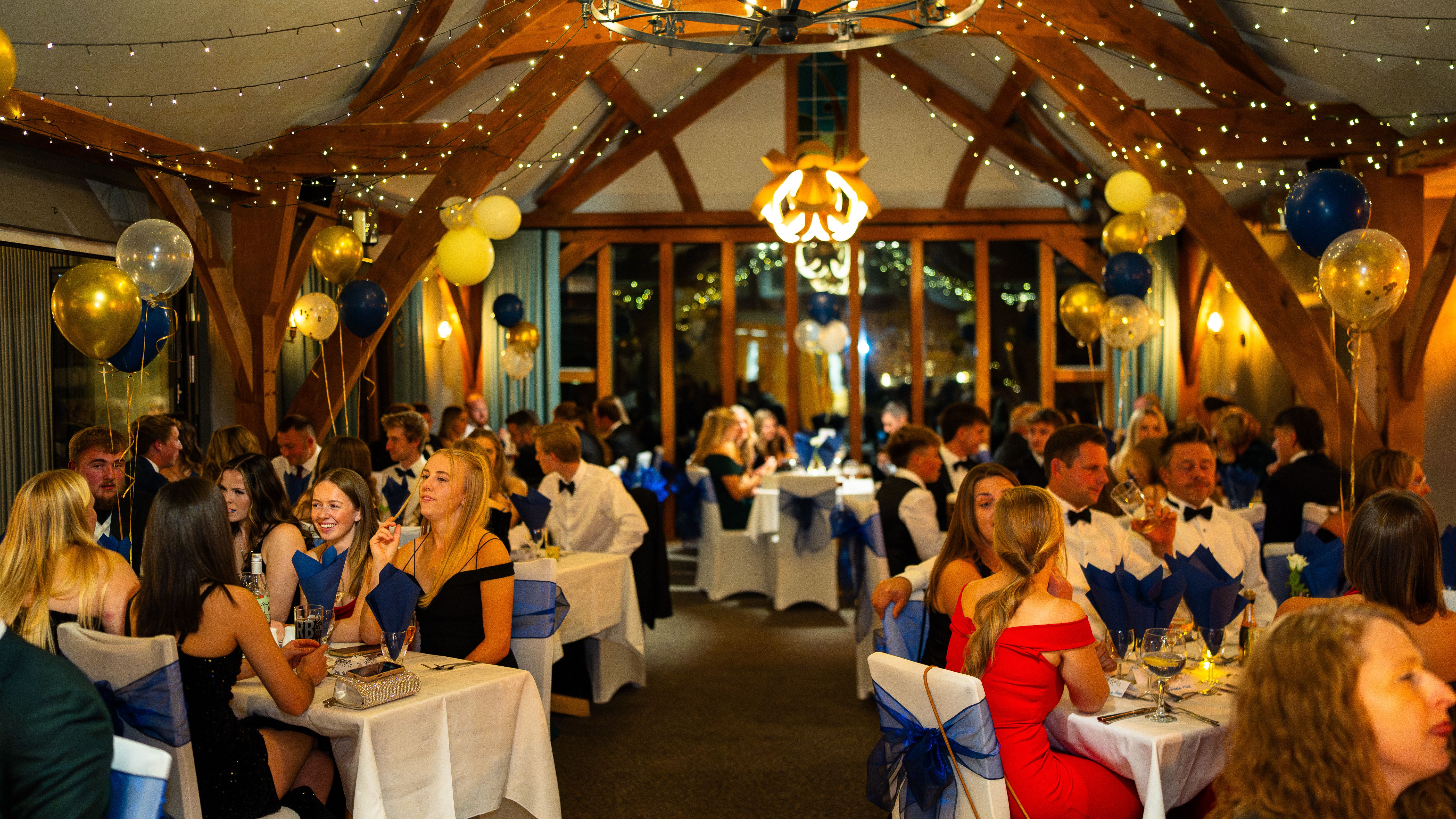 Elegant formal dinner event with people dressed up, sitting at decorated tables with blue and gold balloons and fairy lights.