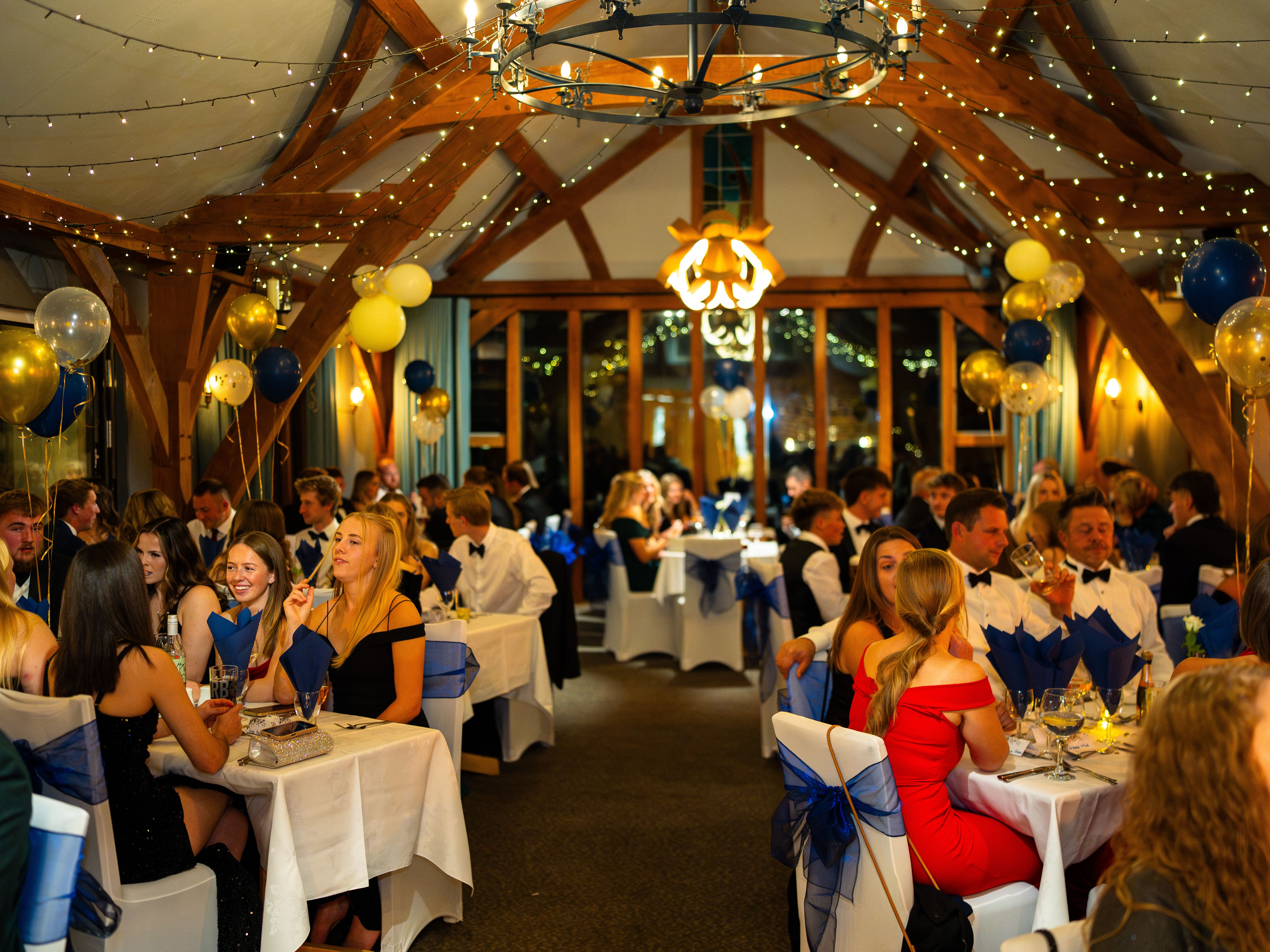 Elegant formal dinner event with people dressed up, sitting at decorated tables with blue and gold balloons and fairy lights.
