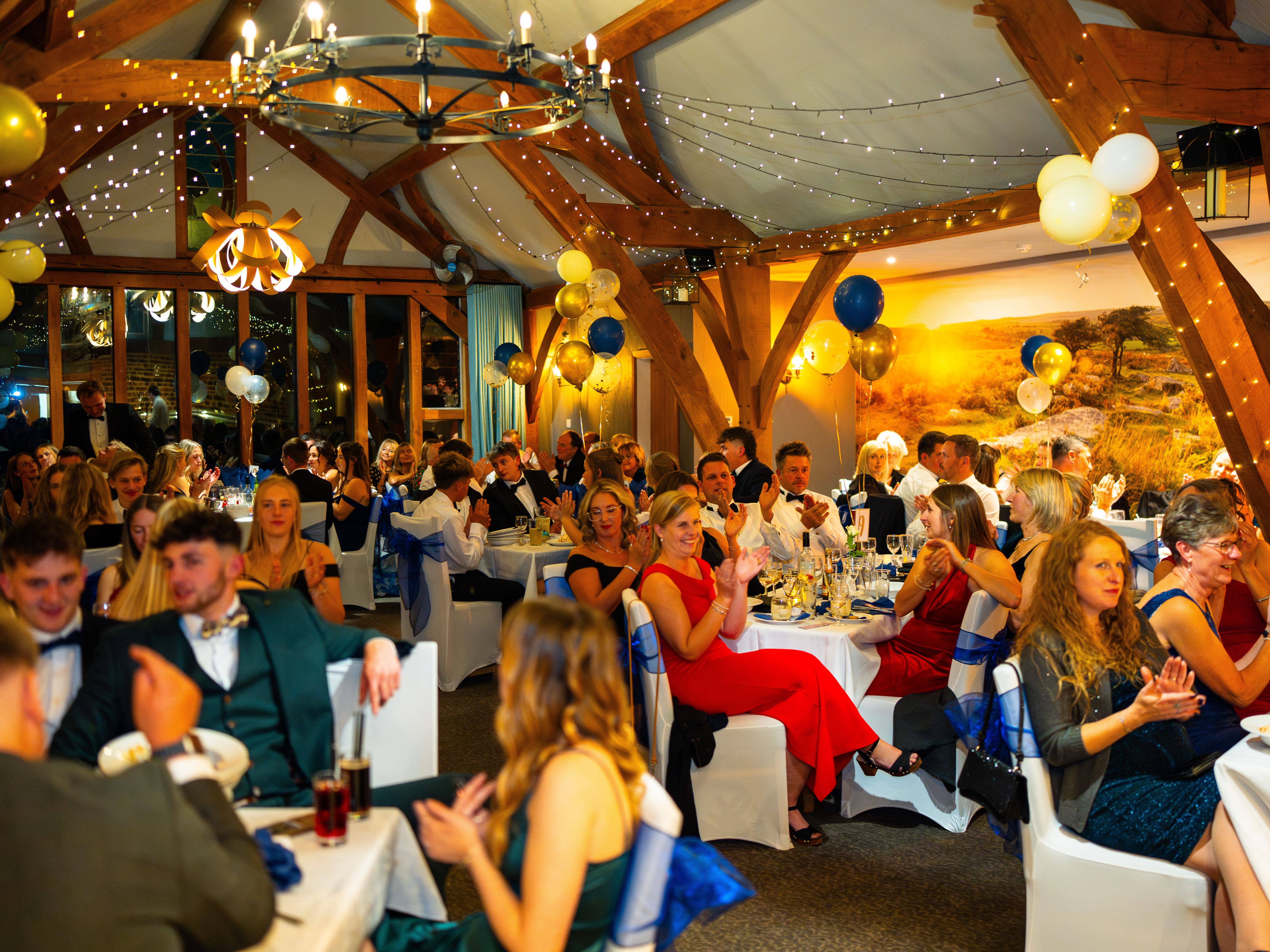 People dressed formally seated at decorated tables during a festive indoor event with balloons and string lights.