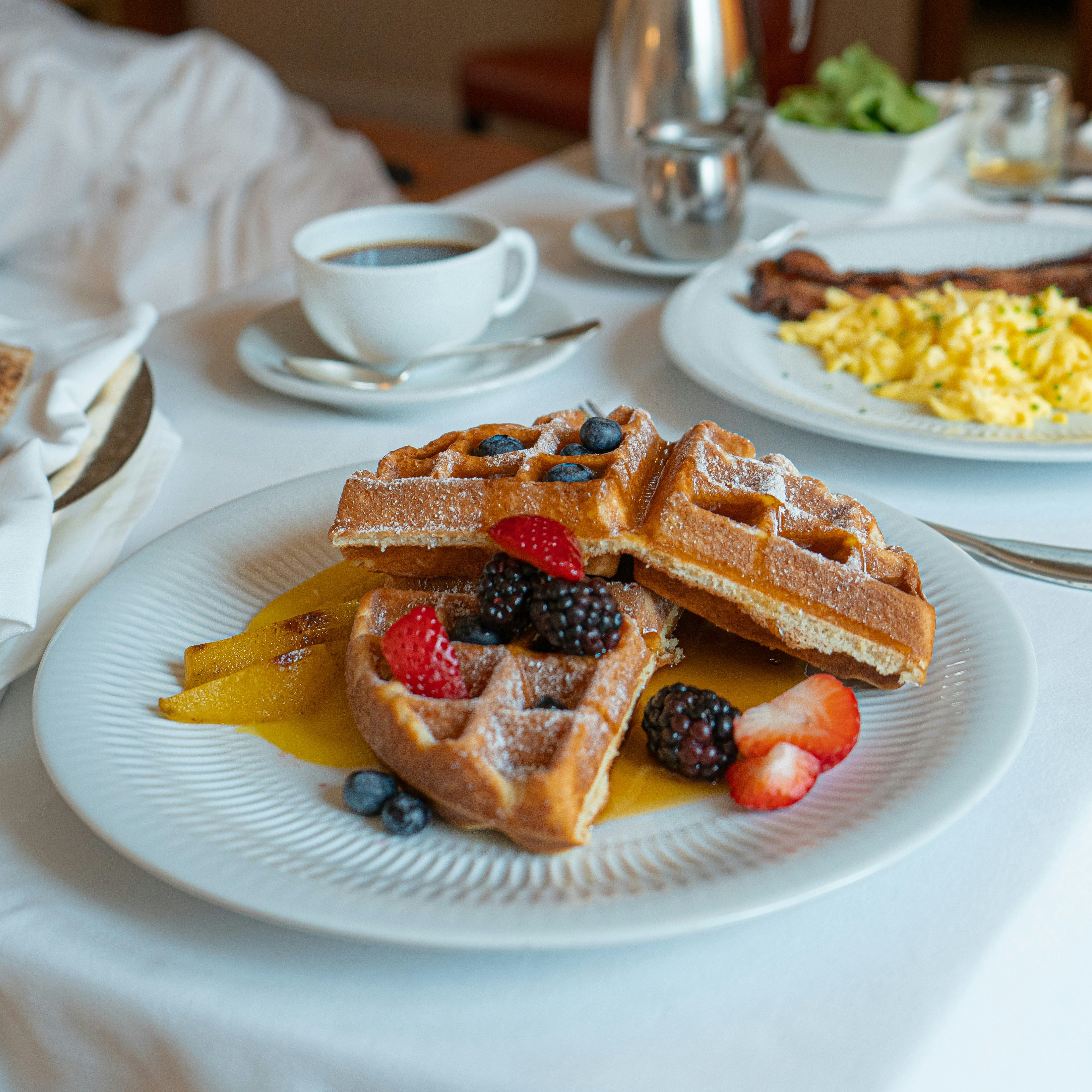 A plate of waffles topped with fresh berries and powdered sugar, served with coffee and breakfast sides on a white tablecloth.