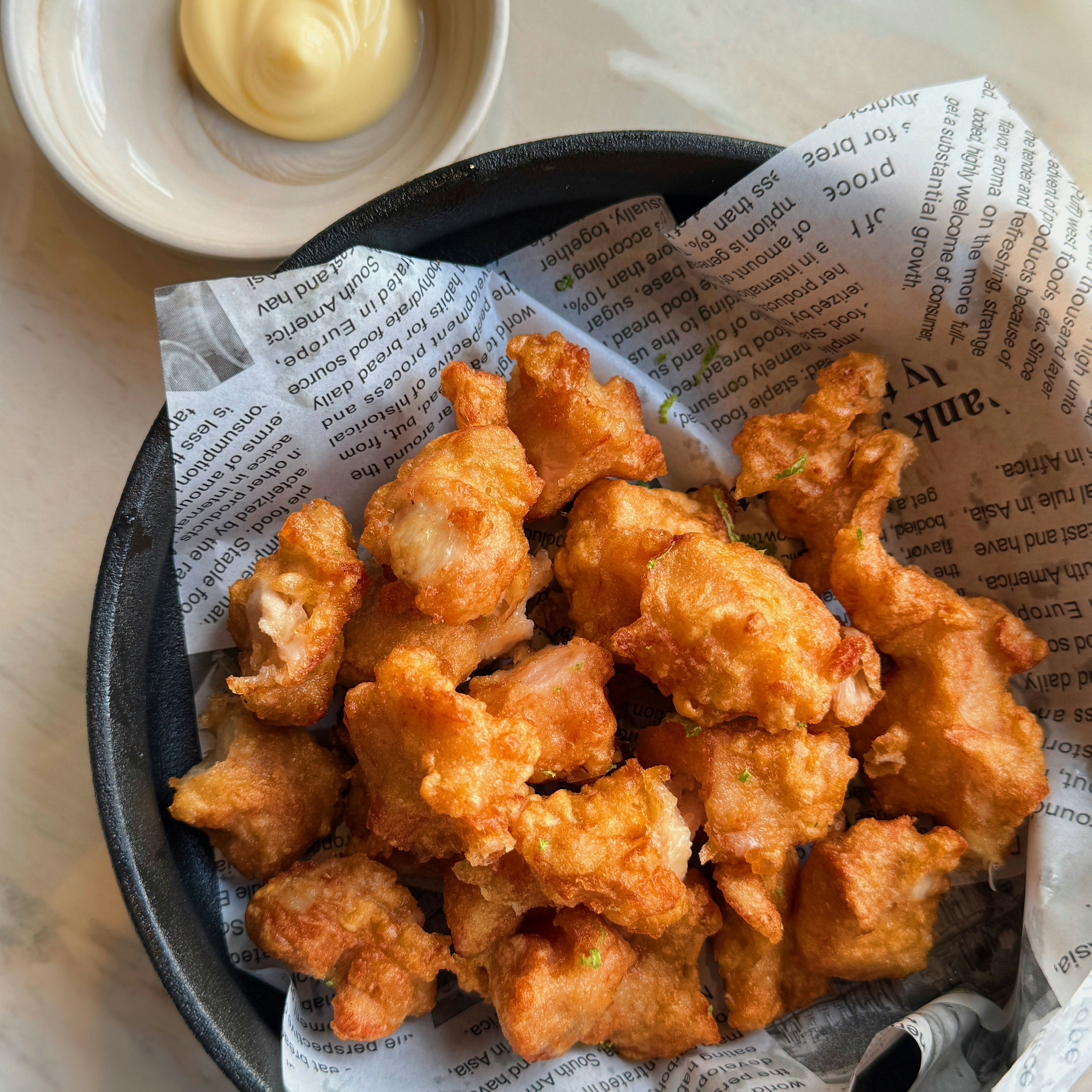 A bowl of golden, crispy fried chicken pieces on newspaper-style paper with a side of creamy mayonnaise dip.