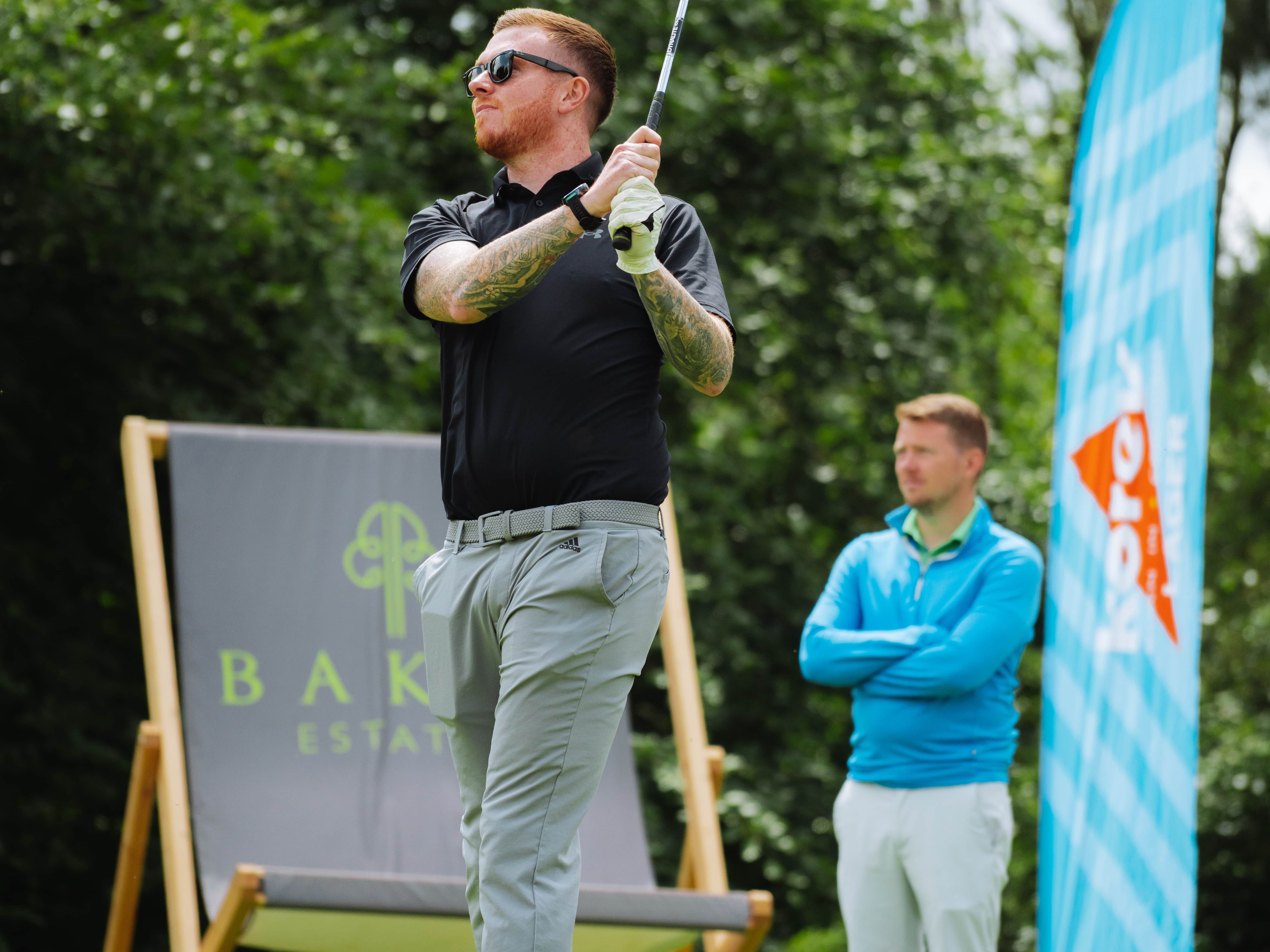 Man swinging a golf club on a green with another man watching in the background