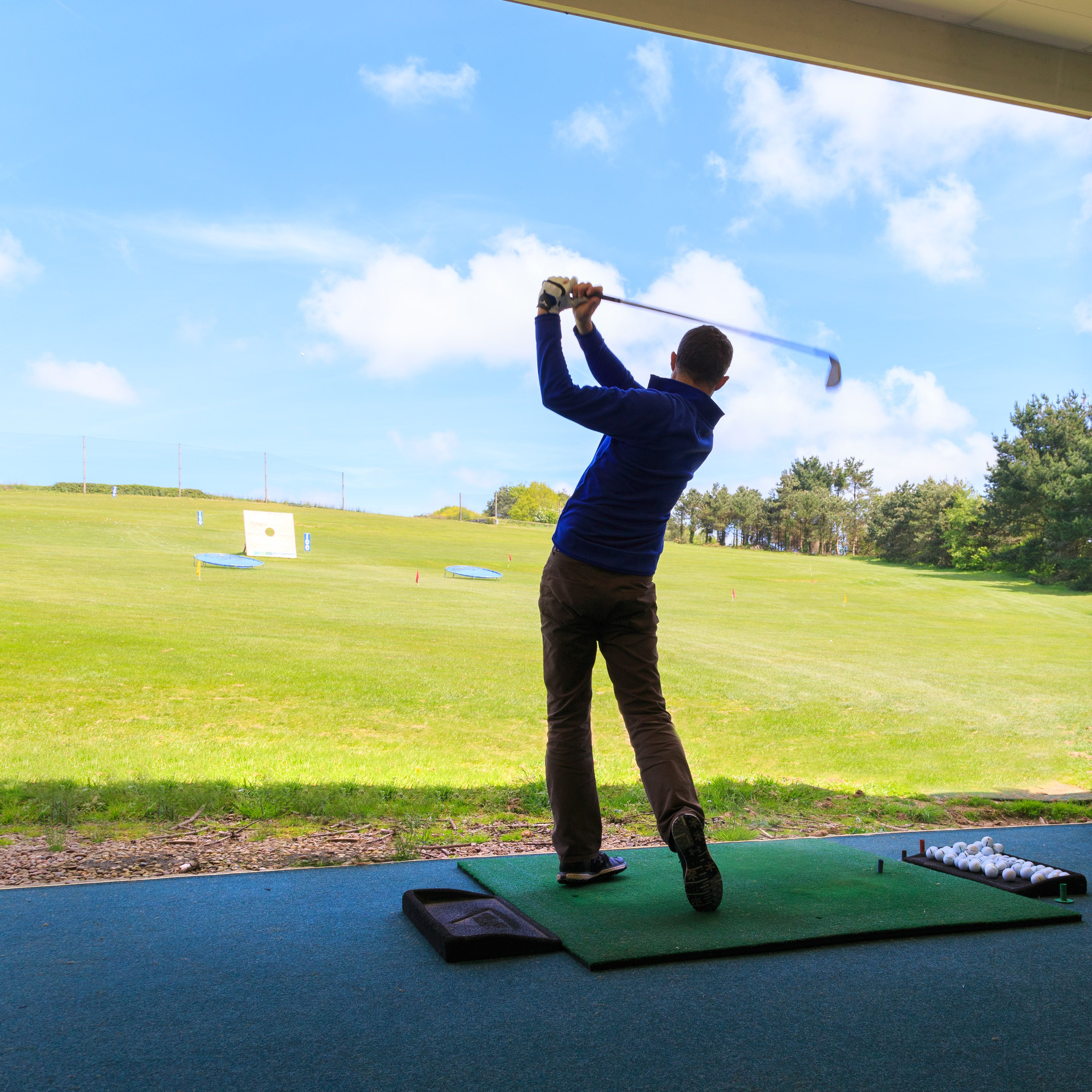 Person practicing golf swing at an indoor driving range bay with another person observing