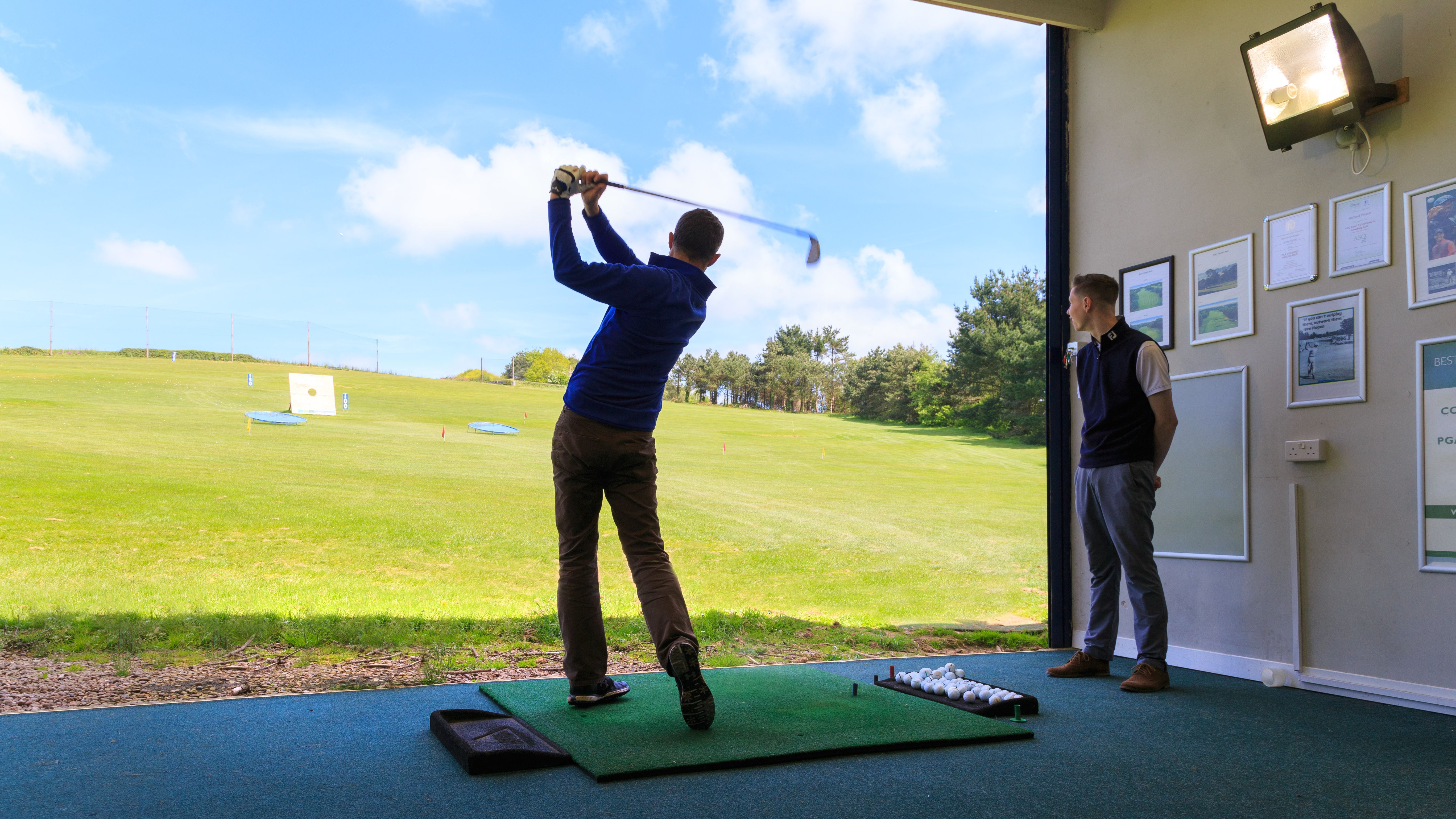 Person practicing golf swing at an indoor driving range bay with another person observing