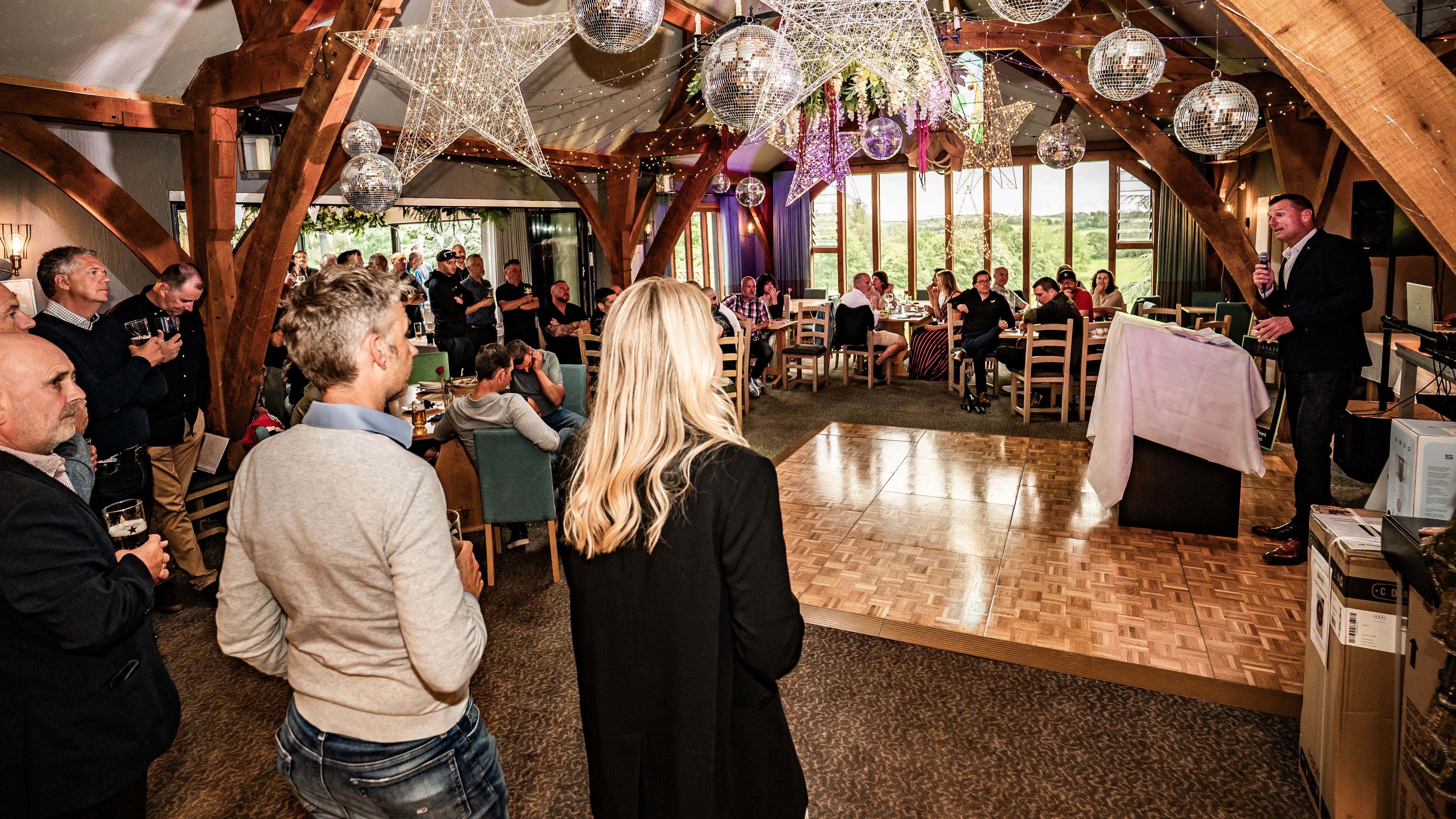 A group of people attending a formal event in a decorated venue, listening to a speaker at a podium.