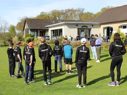 Group of children in black 'Junior Academy' outfits standing on grass near a golf clubhouse, listening to adults.