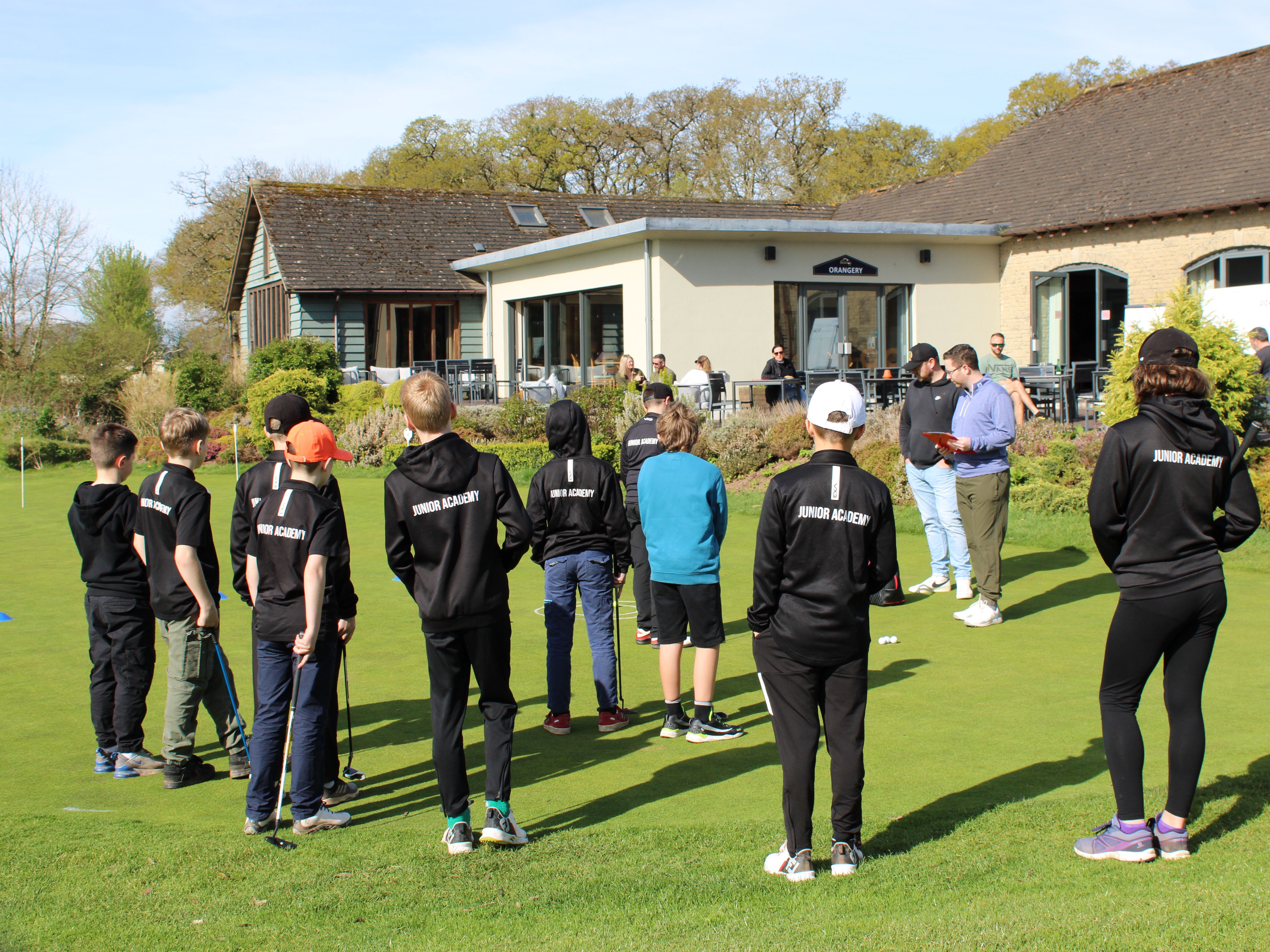 Group of children in black 'Junior Academy' outfits standing on grass near a golf clubhouse, listening to adults.