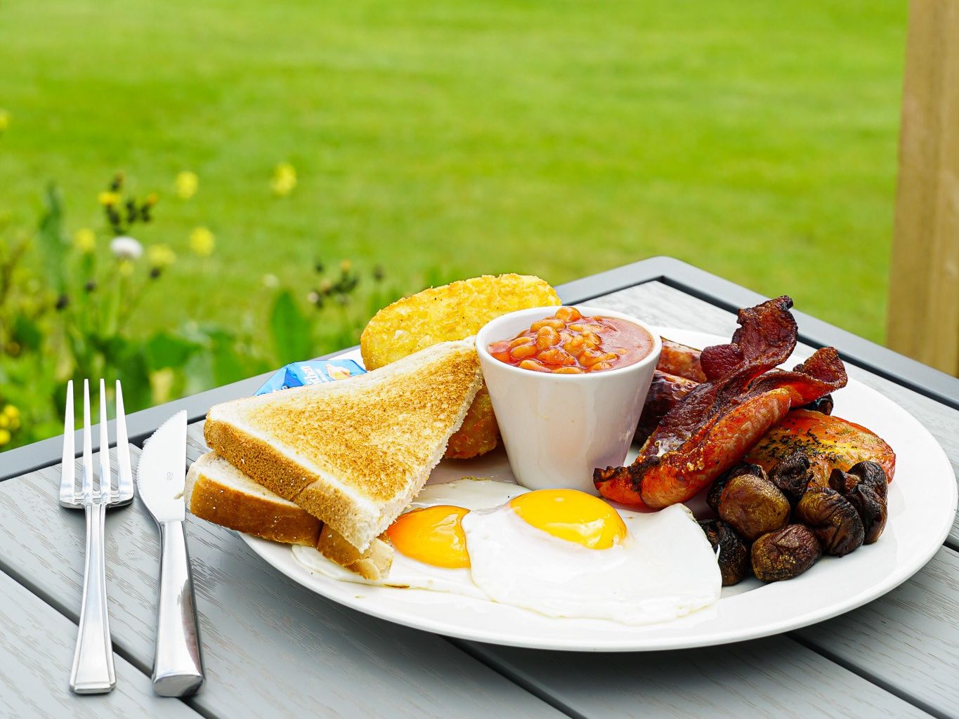Full English breakfast on a plate with eggs, bacon, sausage, beans, toast, hash brown, mushrooms, and tomato, set on a table outdoors with a golf course in the background.
