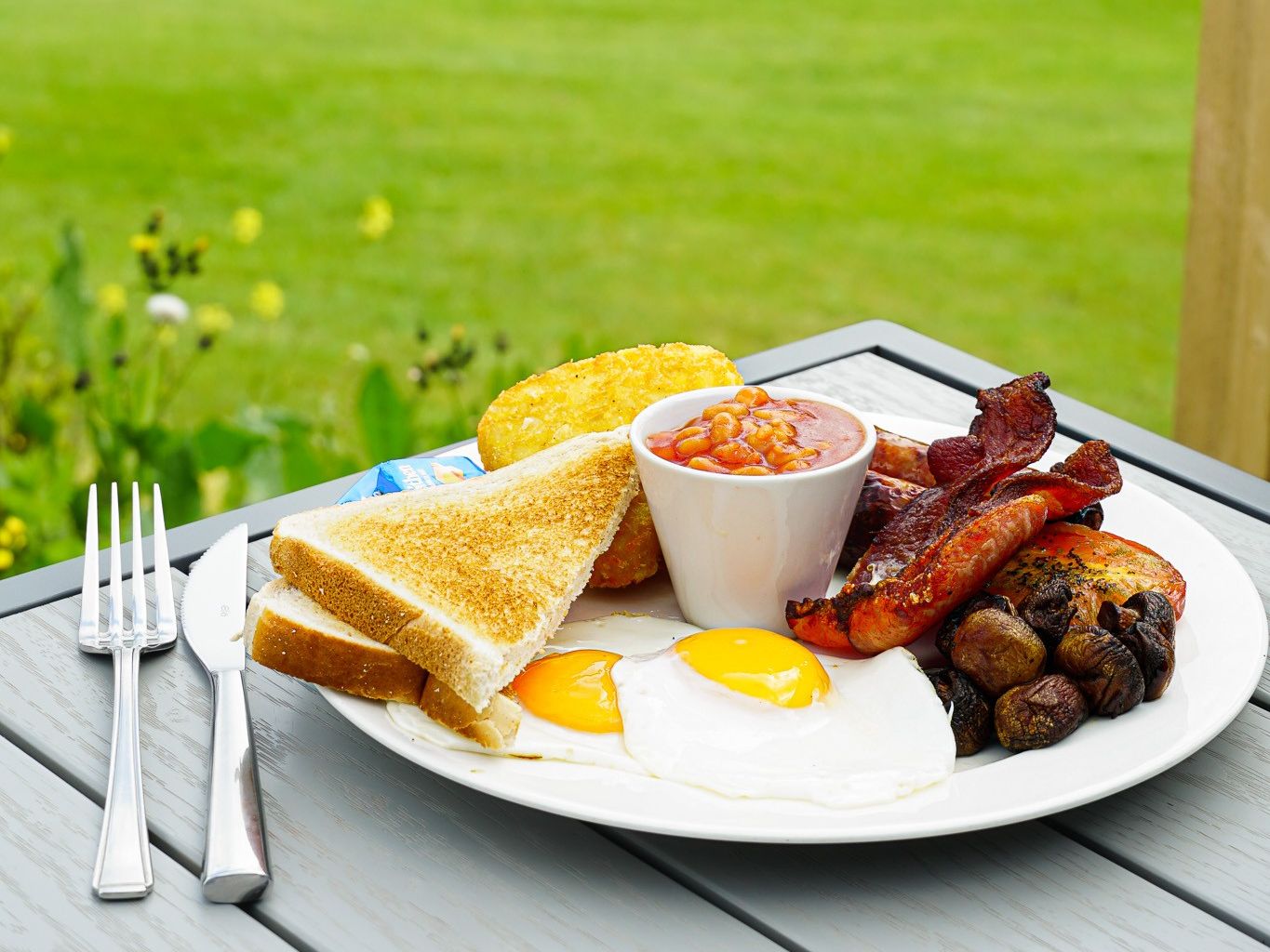 Full English breakfast on a plate with eggs, bacon, sausage, beans, toast, hash brown, mushrooms, and tomato, set on a table outdoors with a golf course in the background.