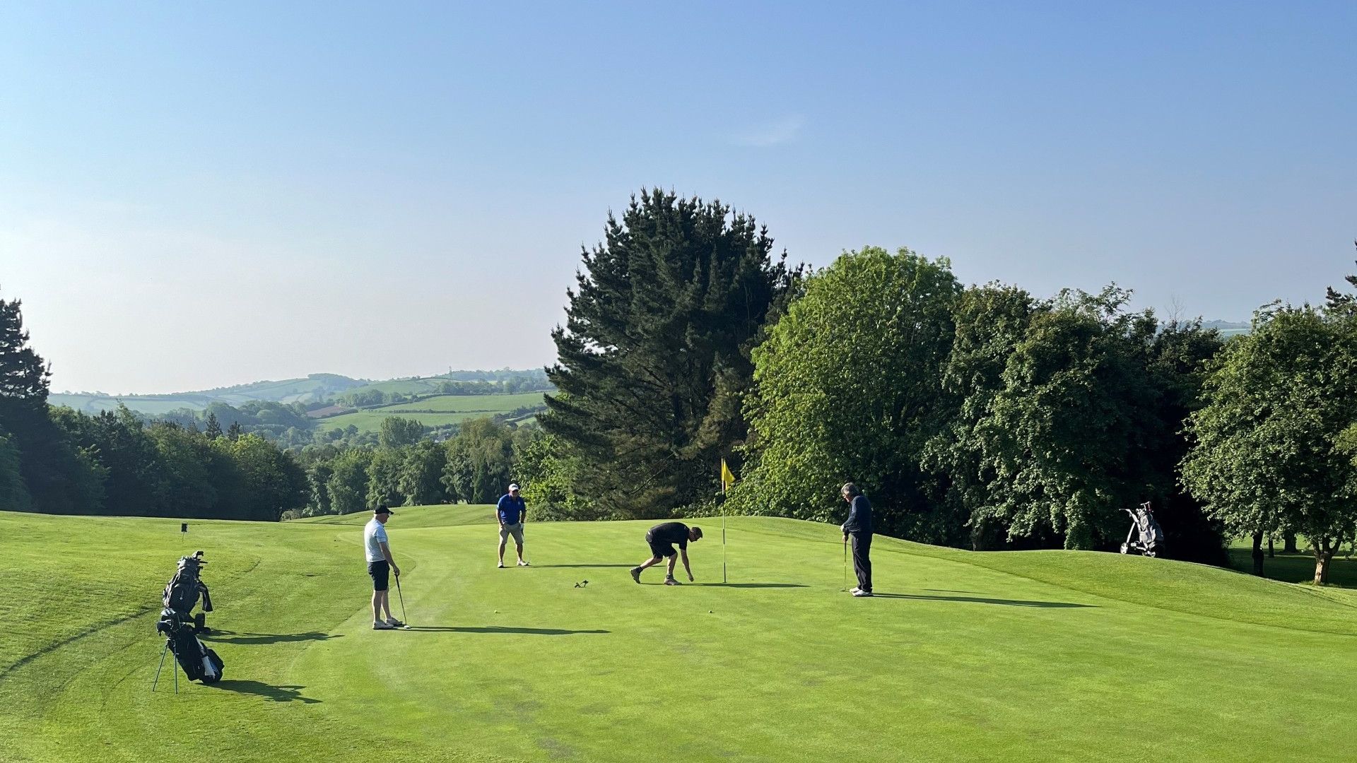Four people playing golf on a green course under a clear blue sky, surrounded by trees and hills.