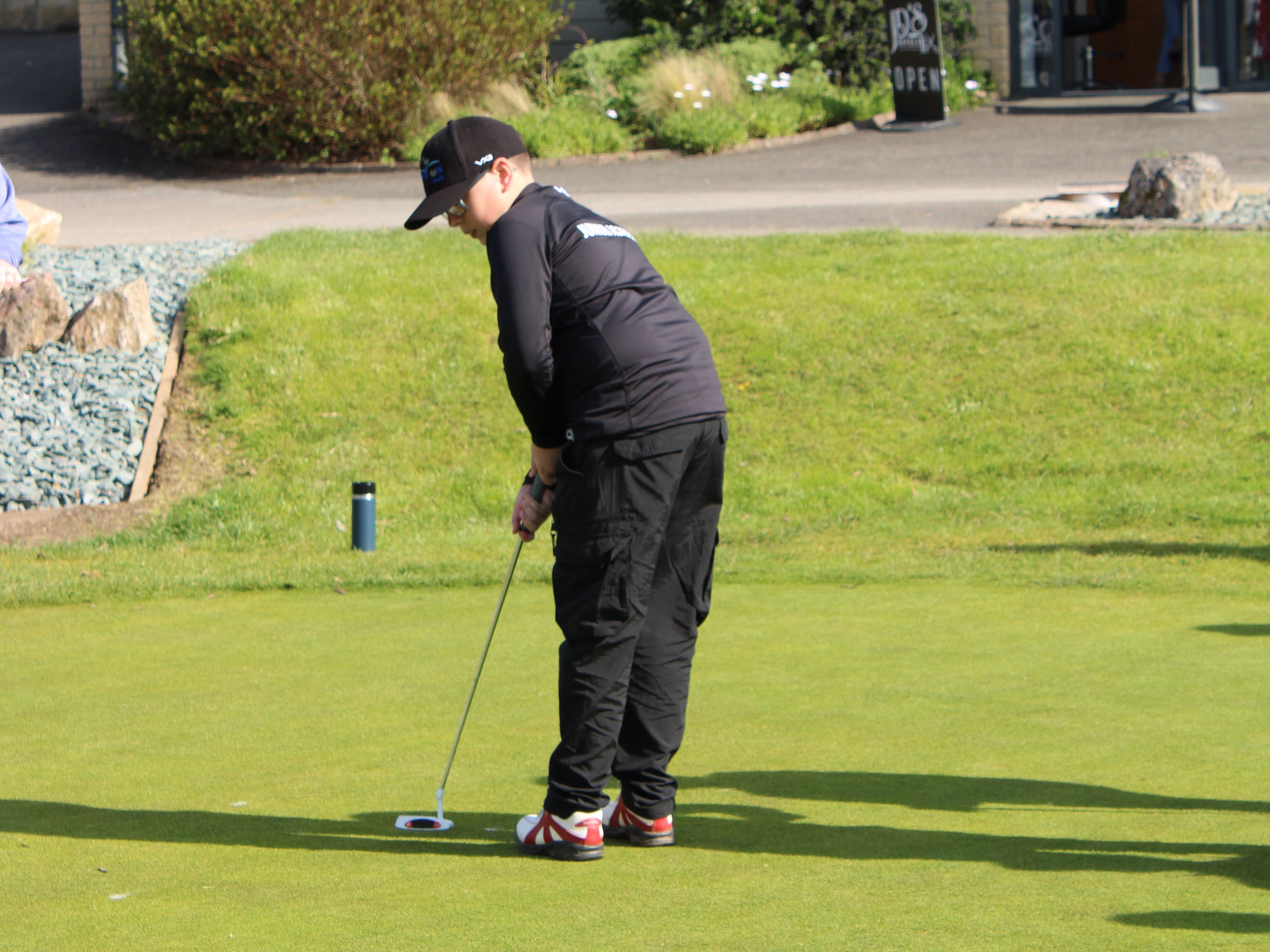 A young person wearing black sportswear and a cap is putting on a golf green near a building with solar panels.