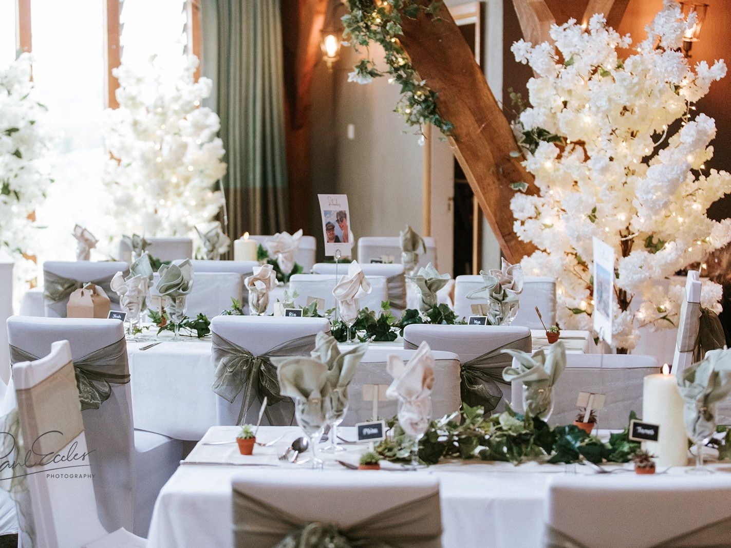 Elegant wedding reception with white and green decor, featuring tables with white cloths, green sashes on chairs, floral centerpieces, and decorative white trees with lights.