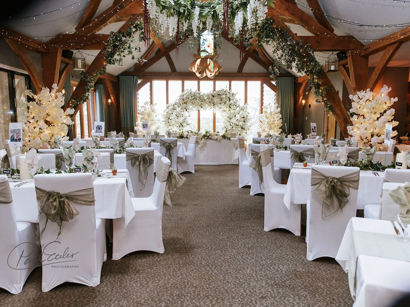 Elegant wedding reception hall decorated with white chair covers, green sashes, floral arrangements, and large windows.