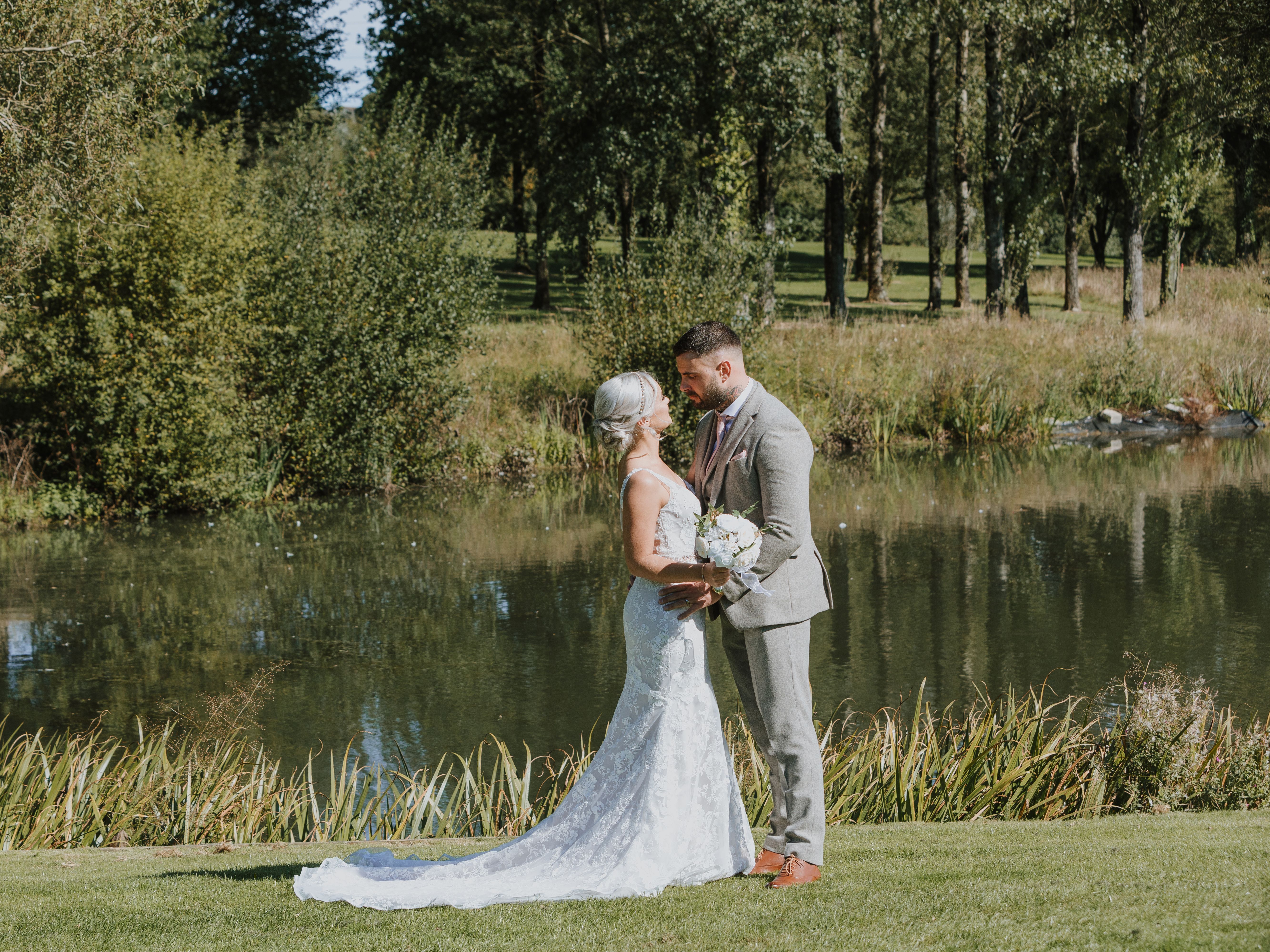 Bride and groom standing close together by a pond surrounded by greenery