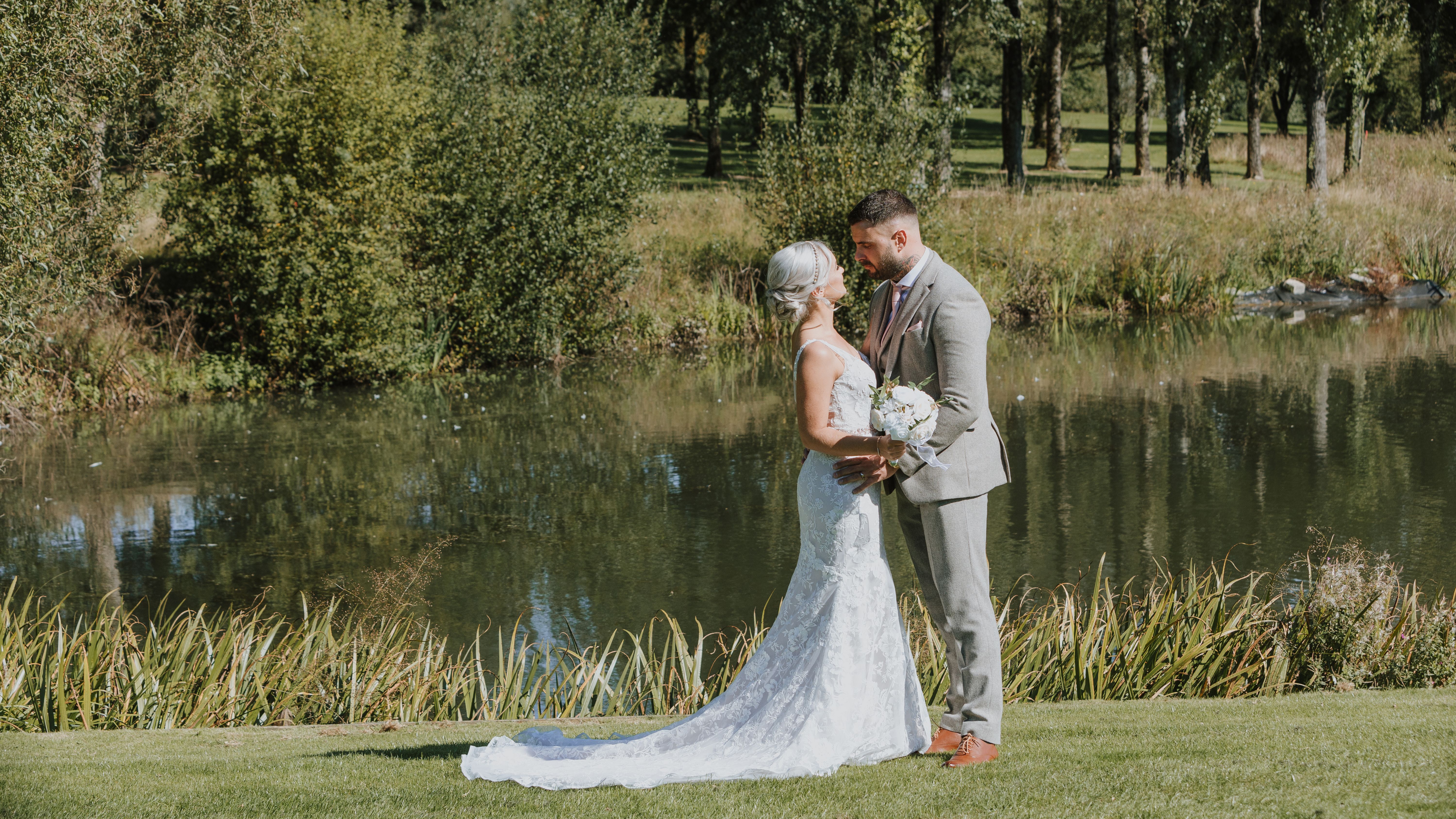 Bride and groom standing close together by a pond surrounded by greenery