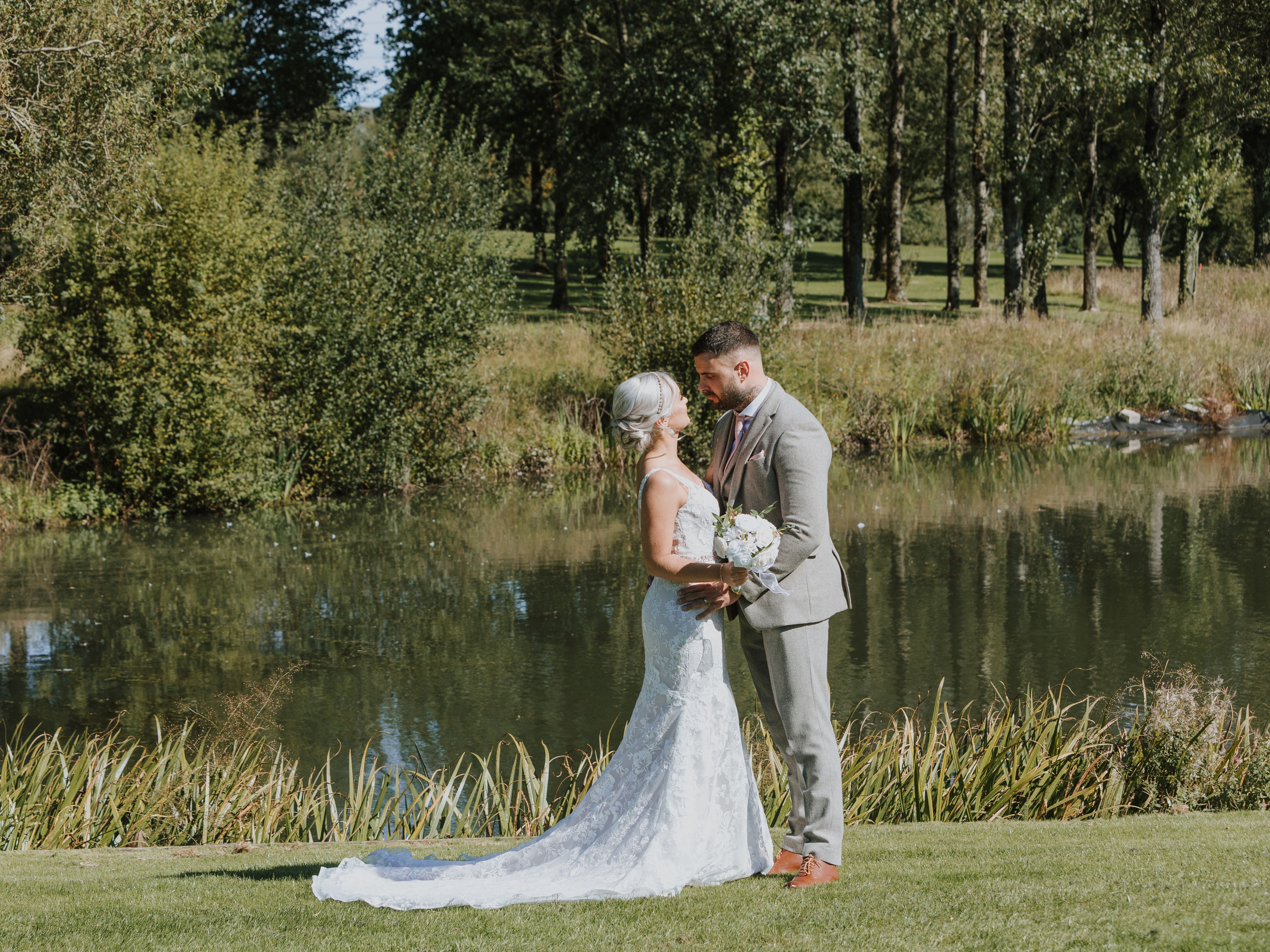Bride and groom standing close together by a pond surrounded by greenery