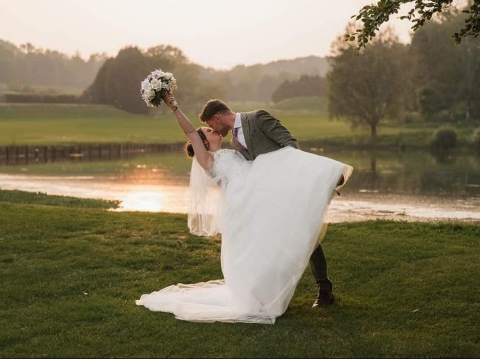 Groom lifts bride in a white wedding dress by a lakeside at sunset, bouquet raised