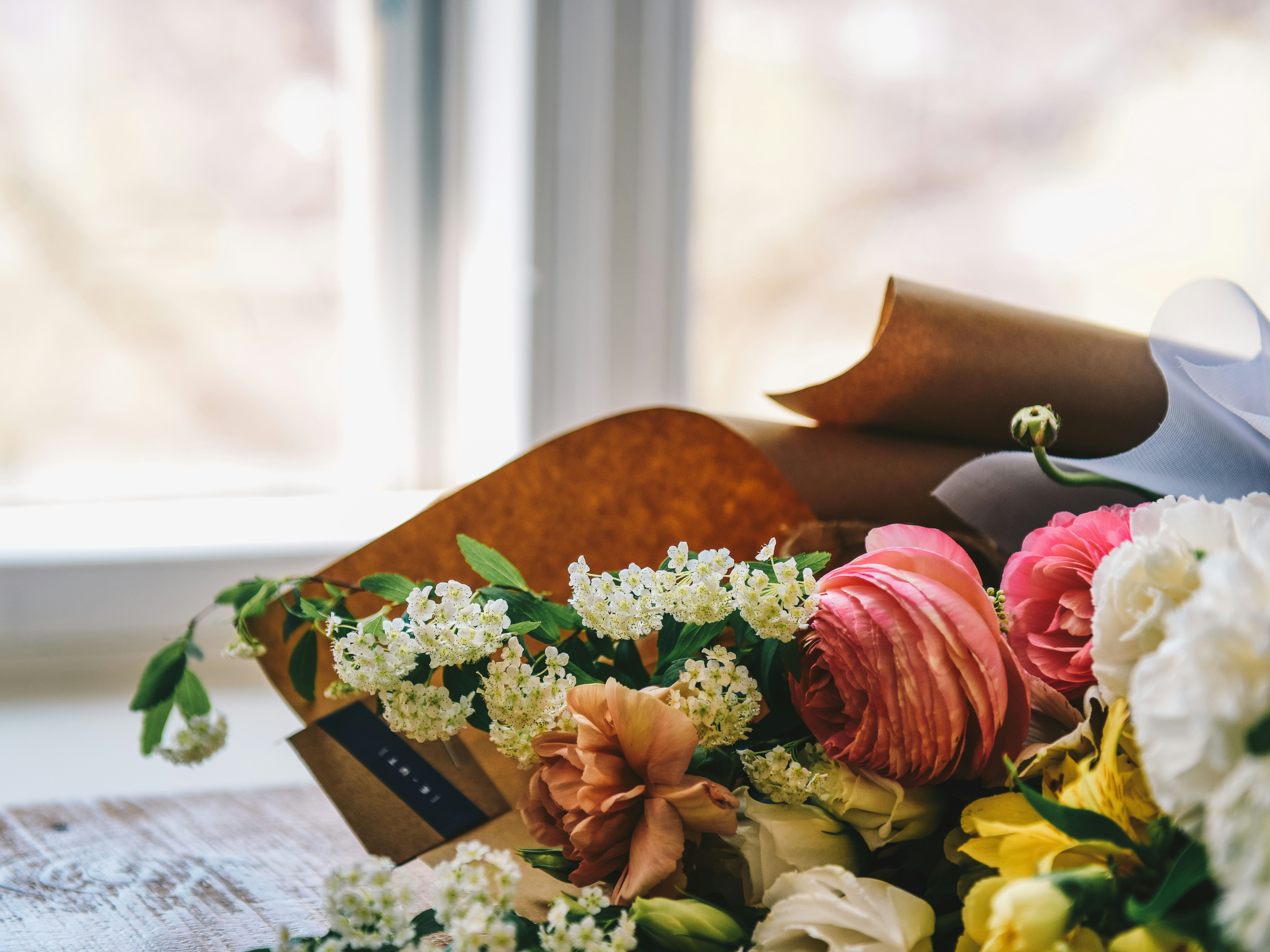 Bouquet of mixed flowers wrapped in brown paper lying on a wooden table by a window