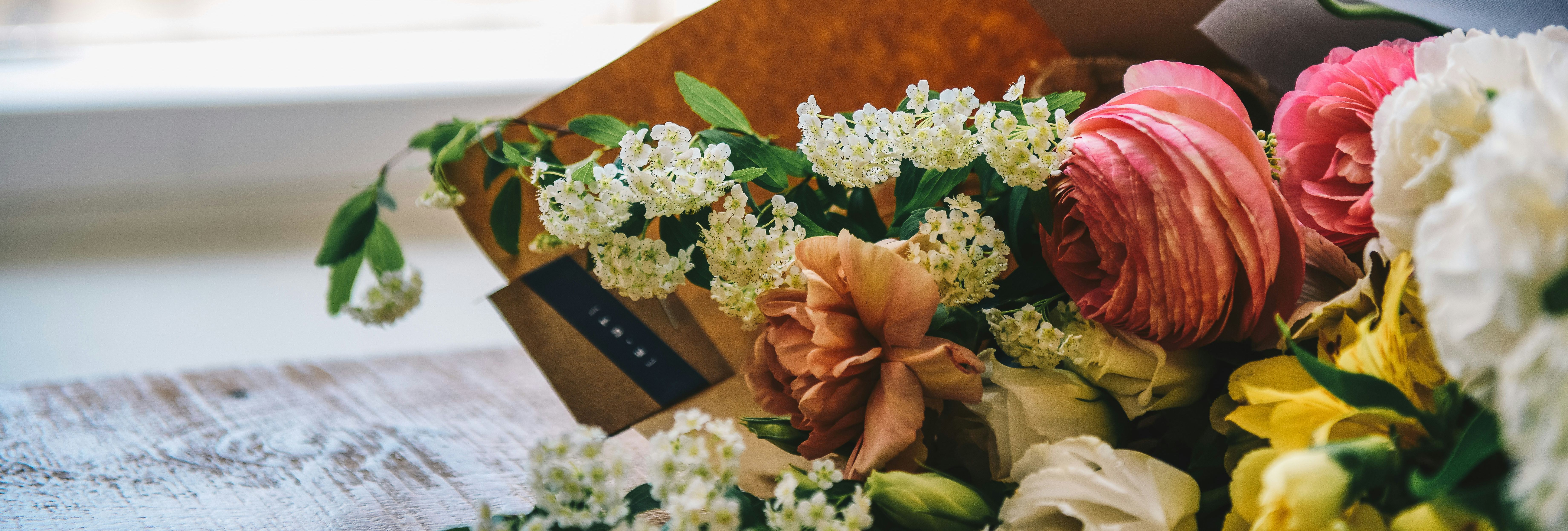 Bouquet of mixed flowers wrapped in brown paper lying on a wooden table by a window