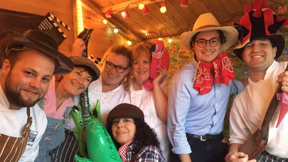Group of colleagues in fancy-dress costumes posing in a wooden hut with string lights, holding inflatable props.