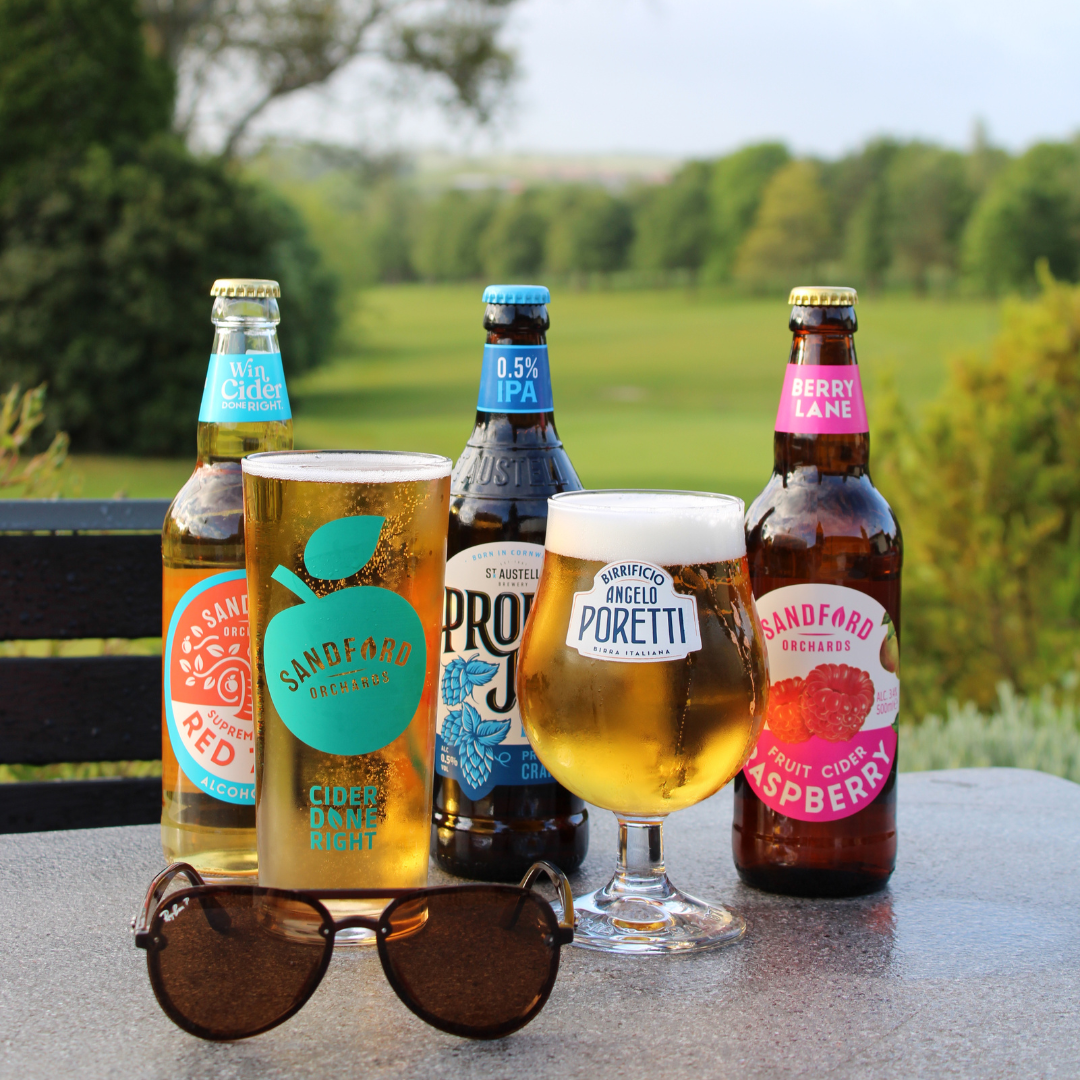 Cider and beer bottles with two glasses on an outdoor table, with sunglasses in front and a green countryside backdrop.