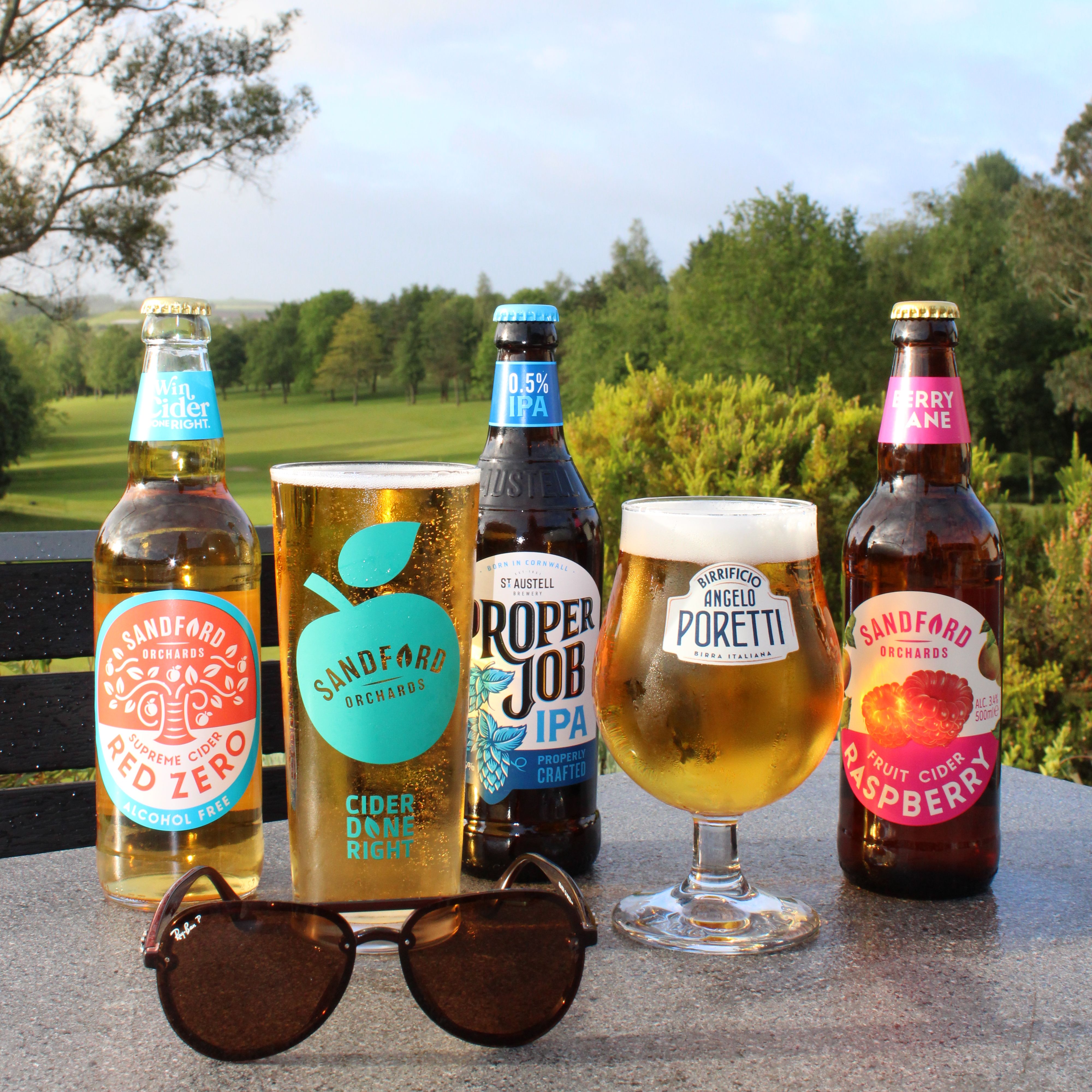 Bottles and glasses of cider and IPA on an outdoor table overlooking a green countryside landscape