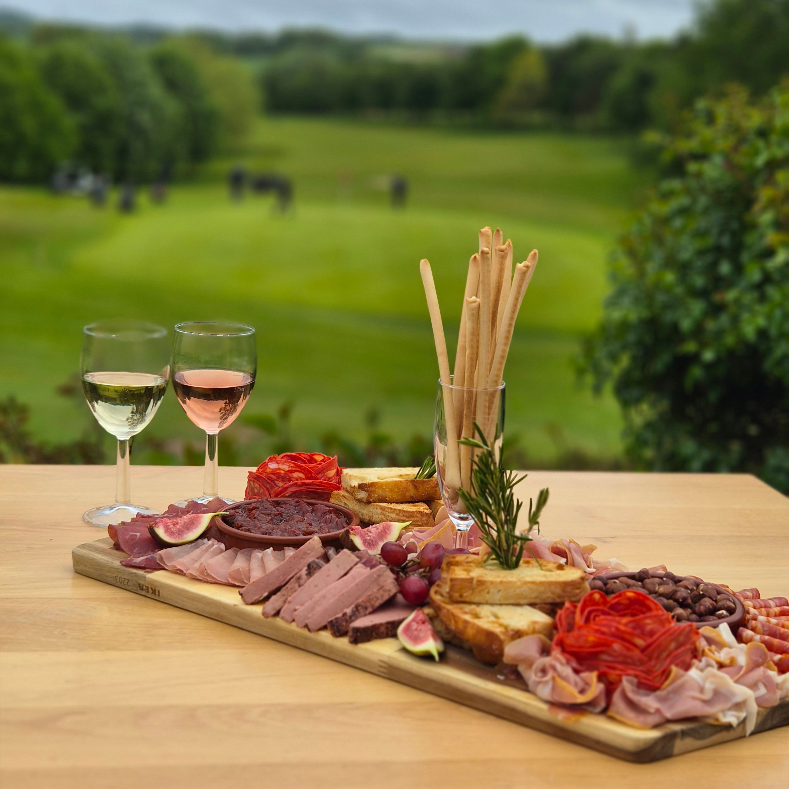 Charcuterie board with cured meats, bread, figs and olives beside two glasses of wine overlooking green countryside