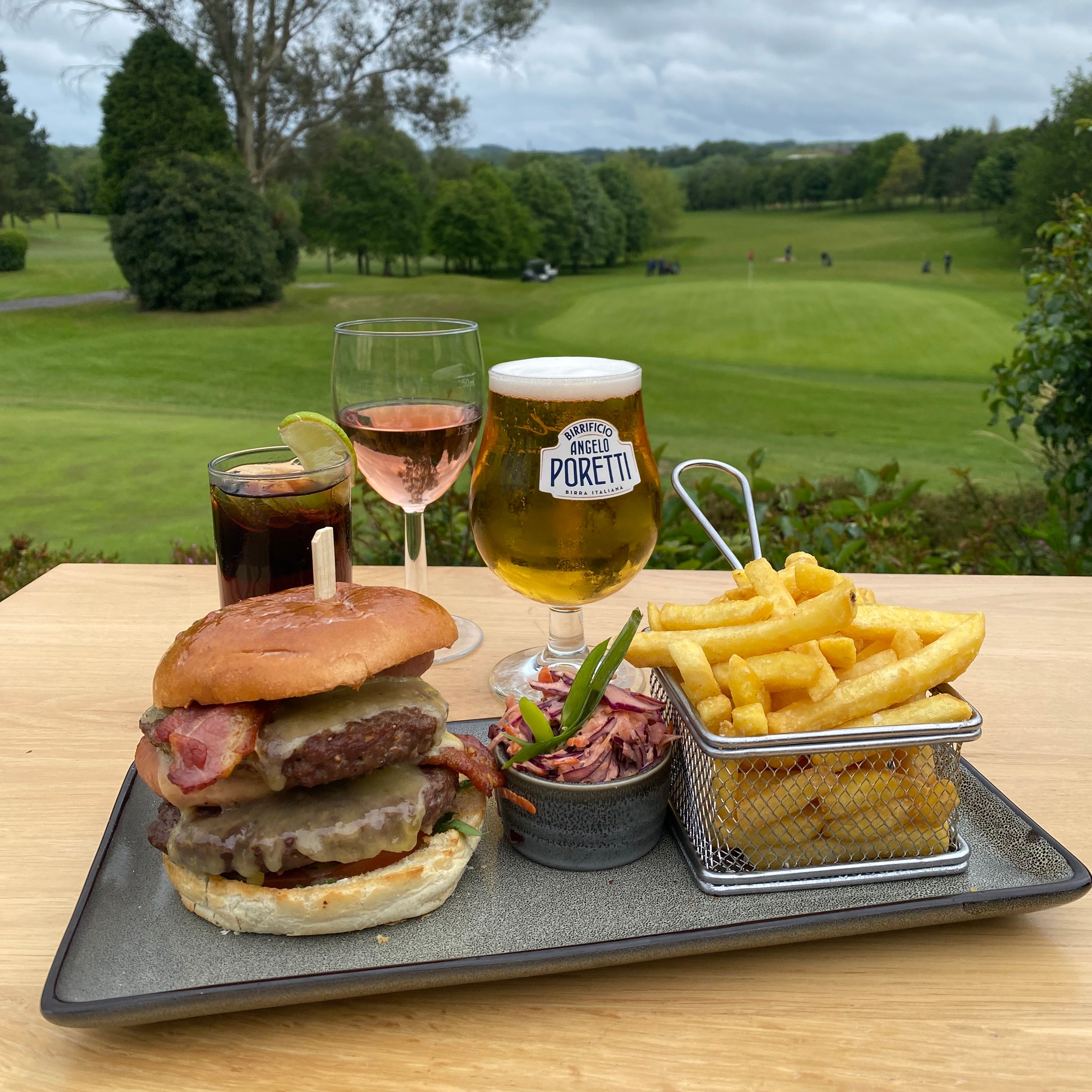 Bacon cheeseburger with chips and slaw on a table, with drinks overlooking a golf course under cloudy skies