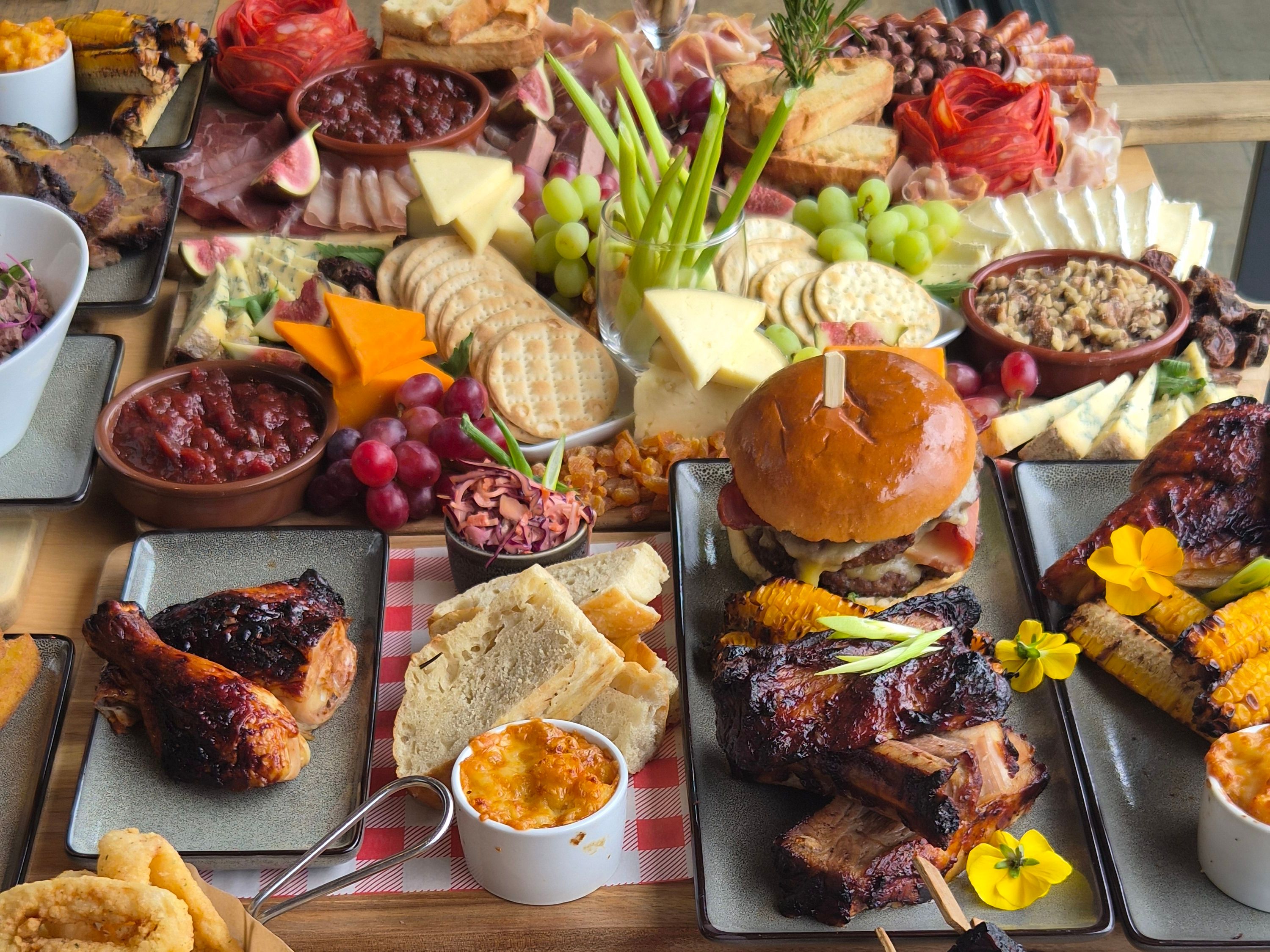Large sharing platter with charcuterie, cheeses, grapes, crackers, toasted bread, burger, ribs and assorted small dishes on a wooden table.