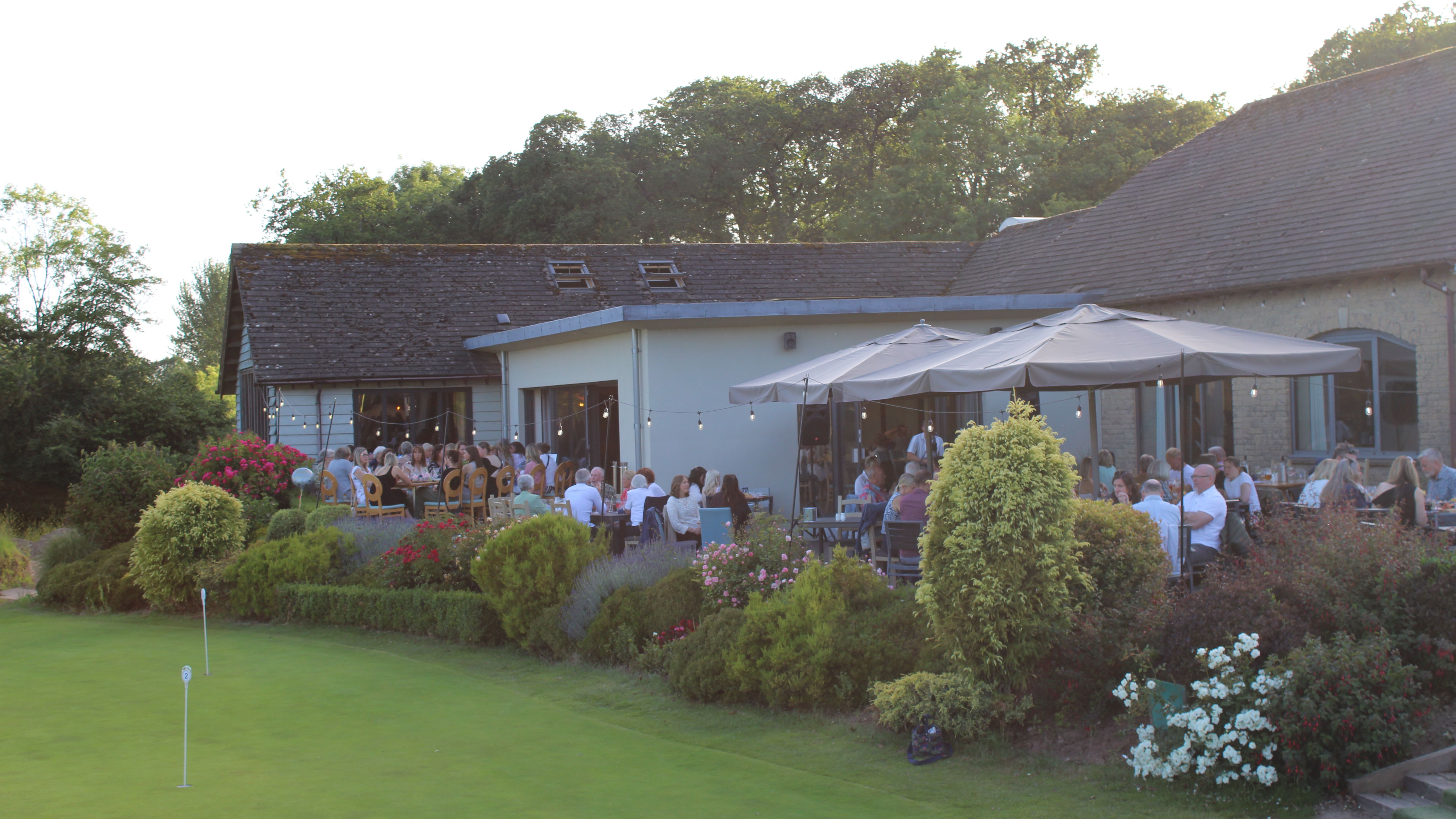 Outdoor terrace dining at a countryside venue beside a putting green, with guests seated under large parasols and string lights
