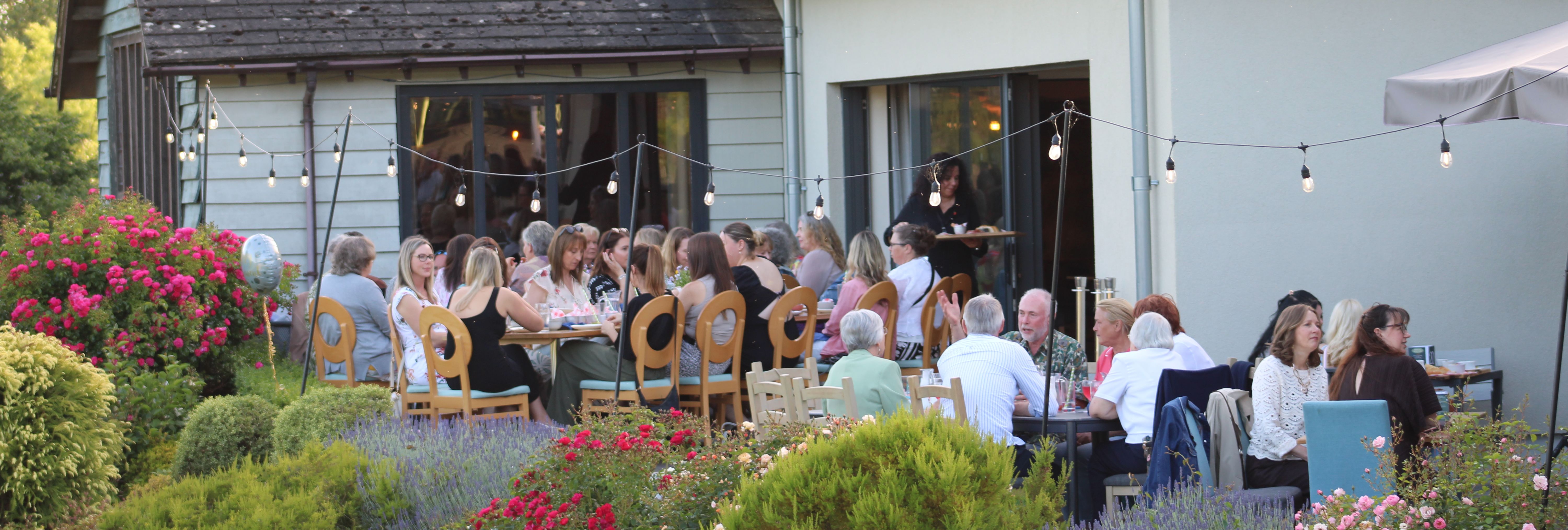 People dining outdoors in a garden beside a modern building with string lights and colourful flowers