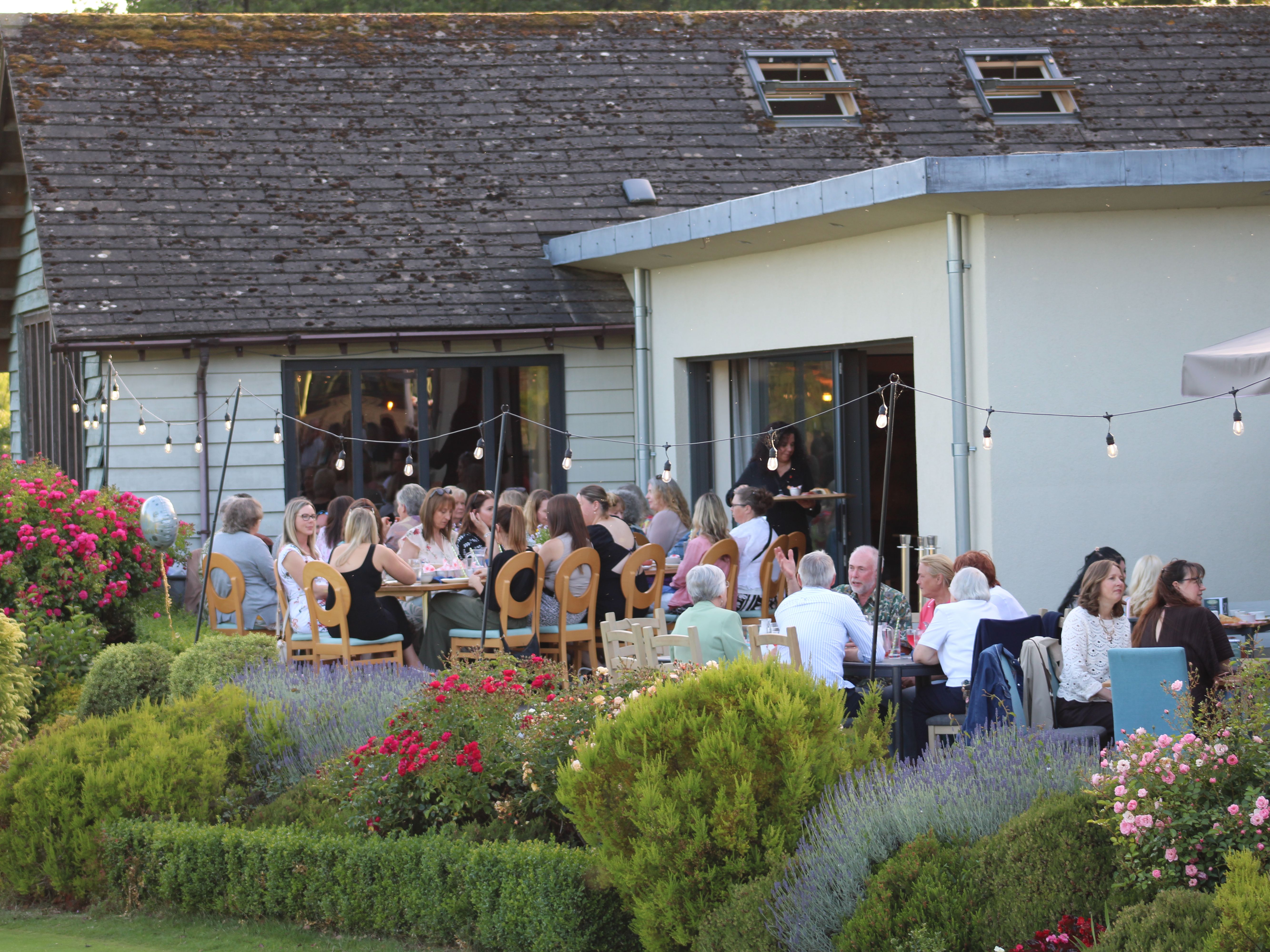 People dining outdoors in a garden beside a modern building with string lights and colourful flowers