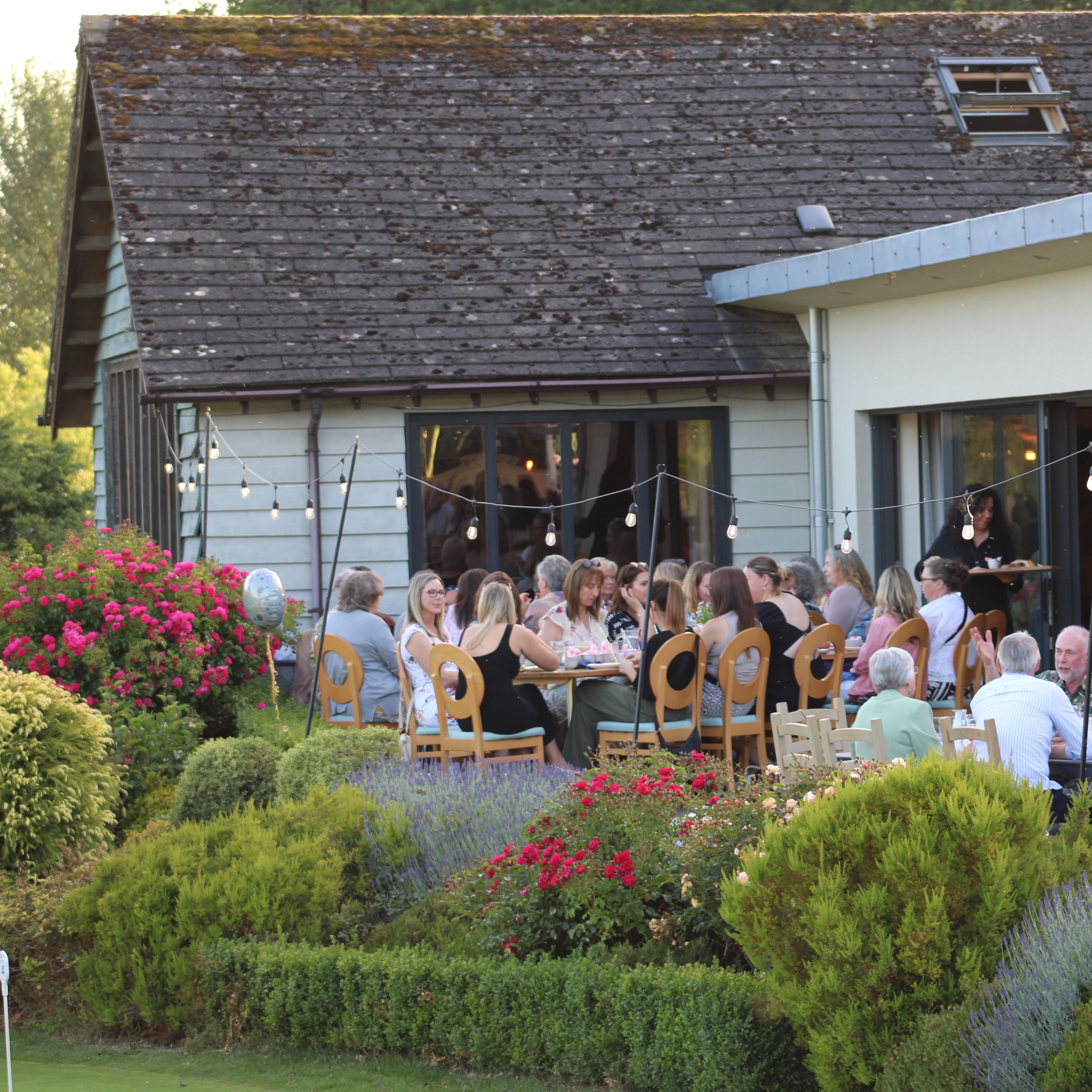 People dining outdoors in a garden beside a modern building with string lights and colourful flowers