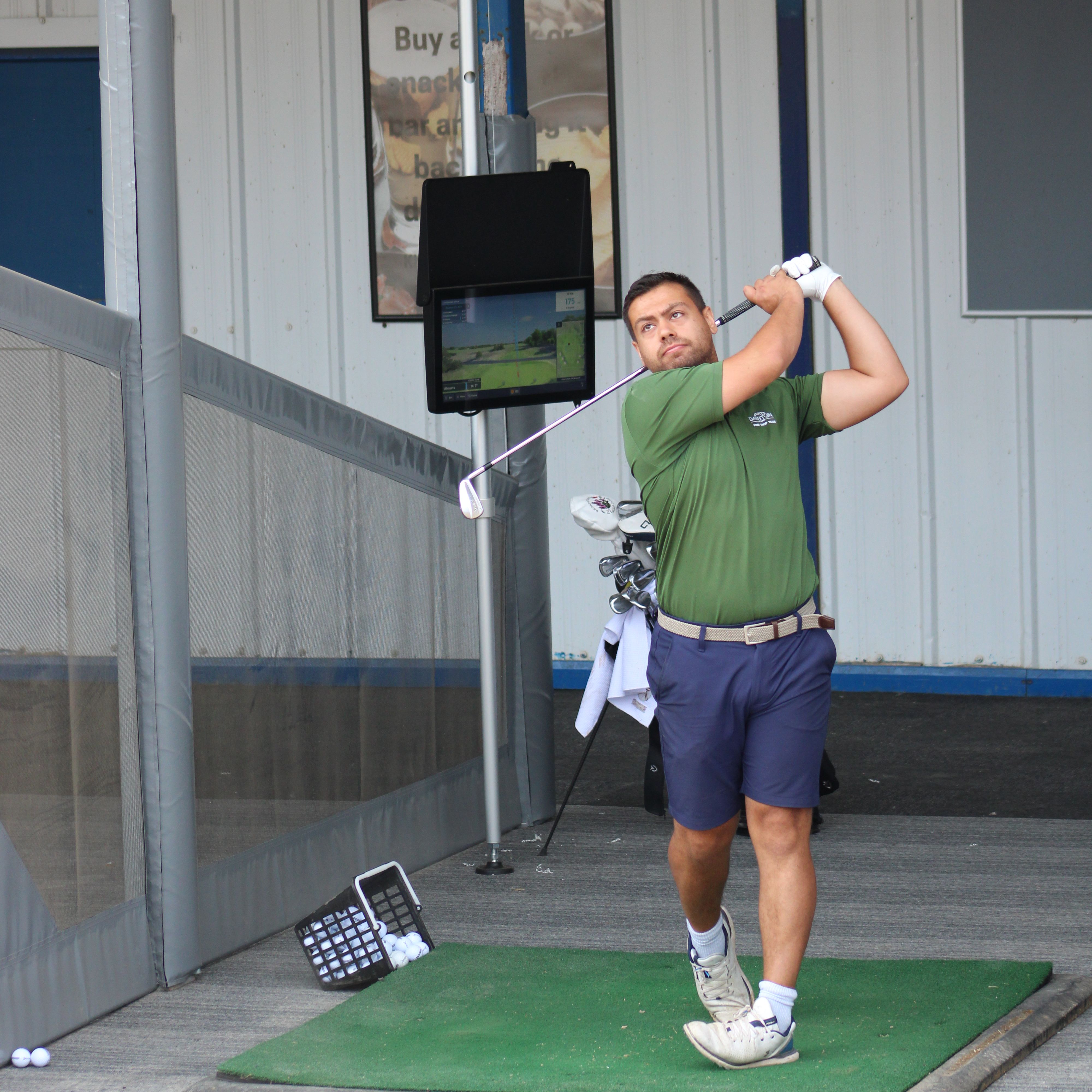 Golfer finishing a swing on a driving range mat beside a launch monitor screen