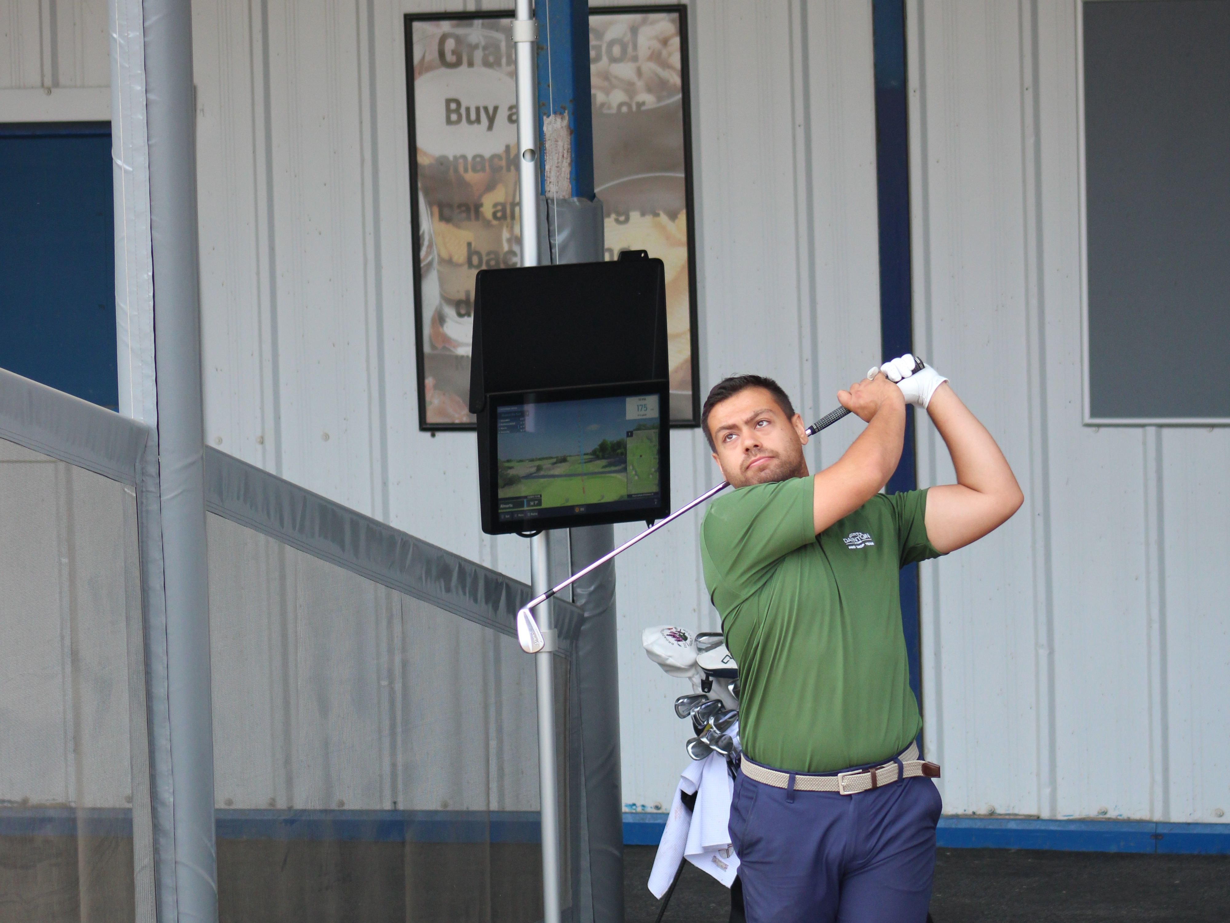 Golfer finishing a swing on a driving range mat beside a launch monitor screen