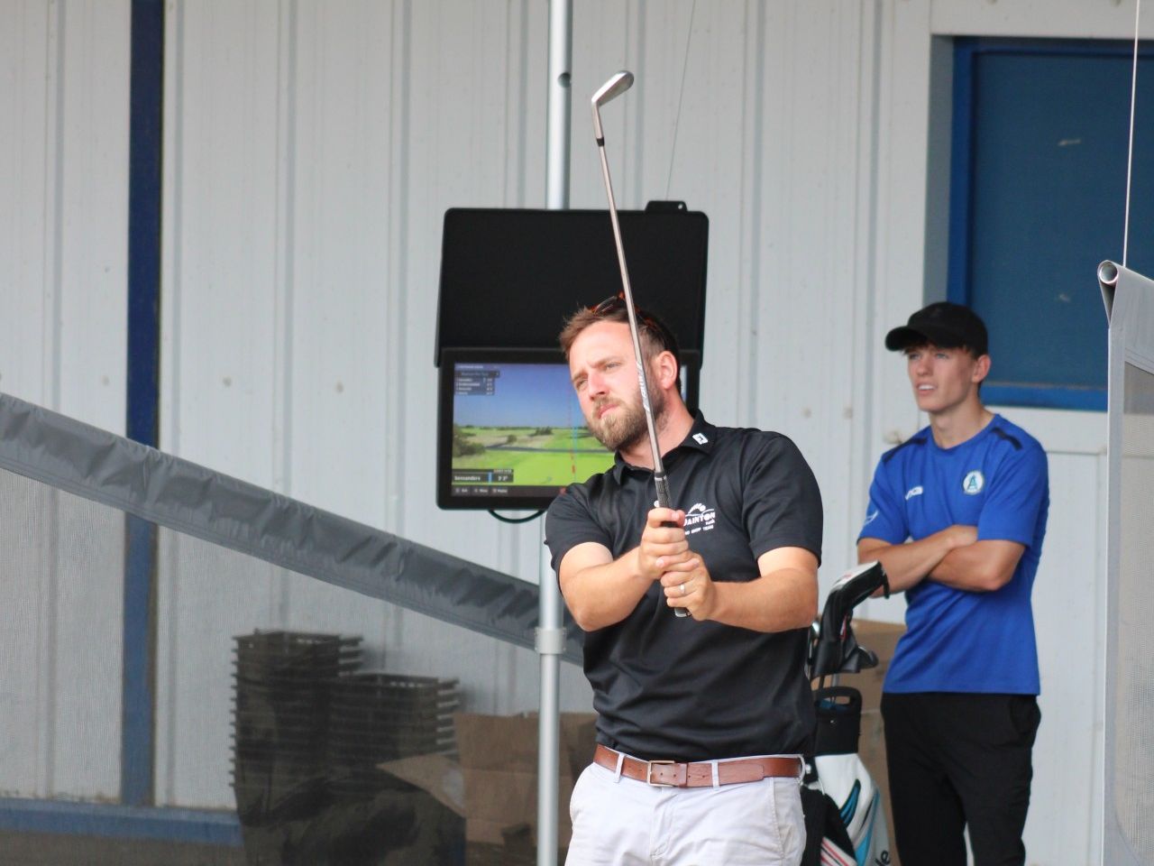 Man practising a golf swing at a driving range bay with a launch monitor screen behind him